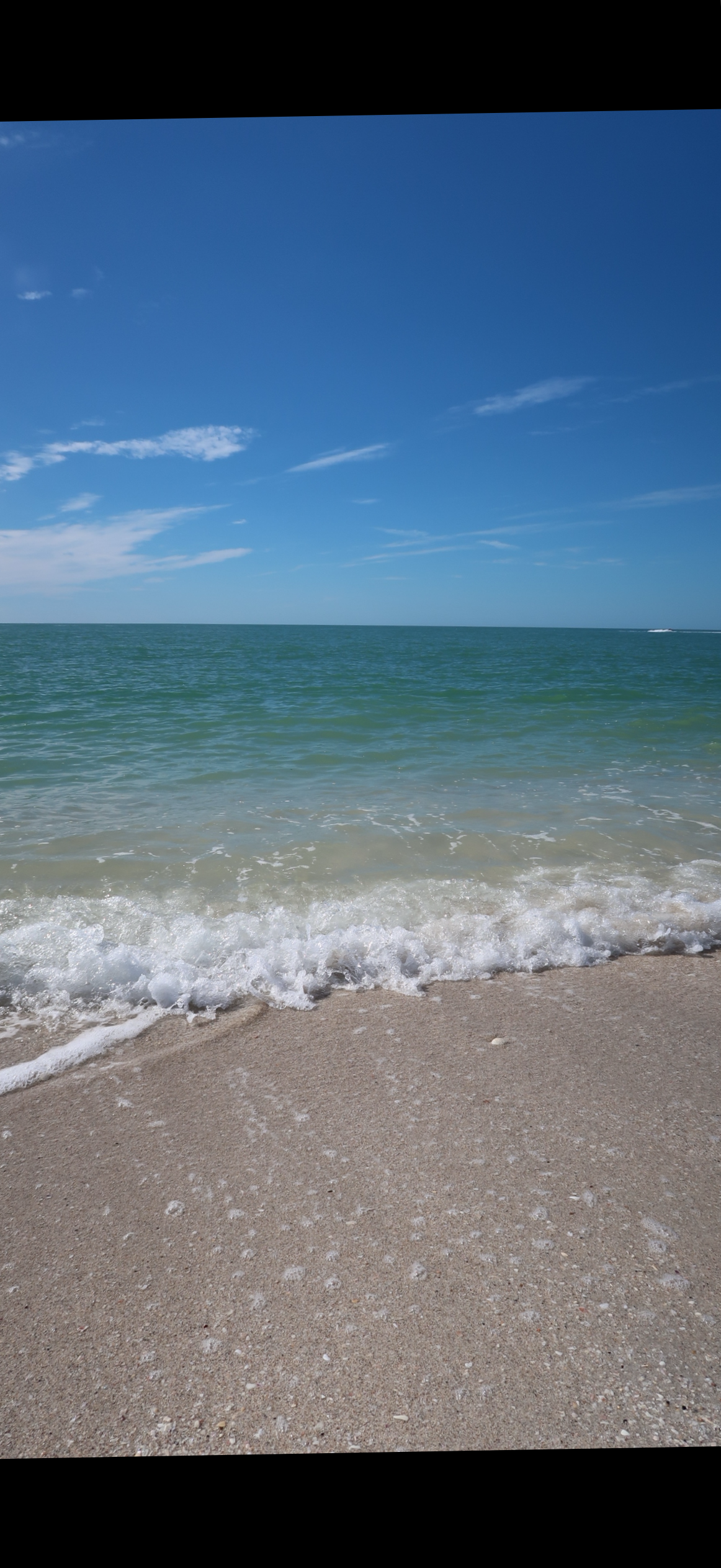A peaceful beach scene with gentle waves lapping against sandy shore, under a clear blue sky with some scattered clouds.