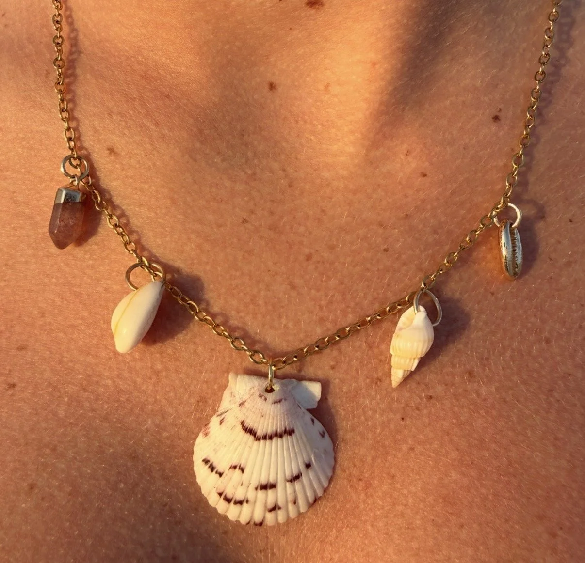A close-up of a person's neck wearing a gold necklace with multiple small shell charms and a larger shell pendant in the center.