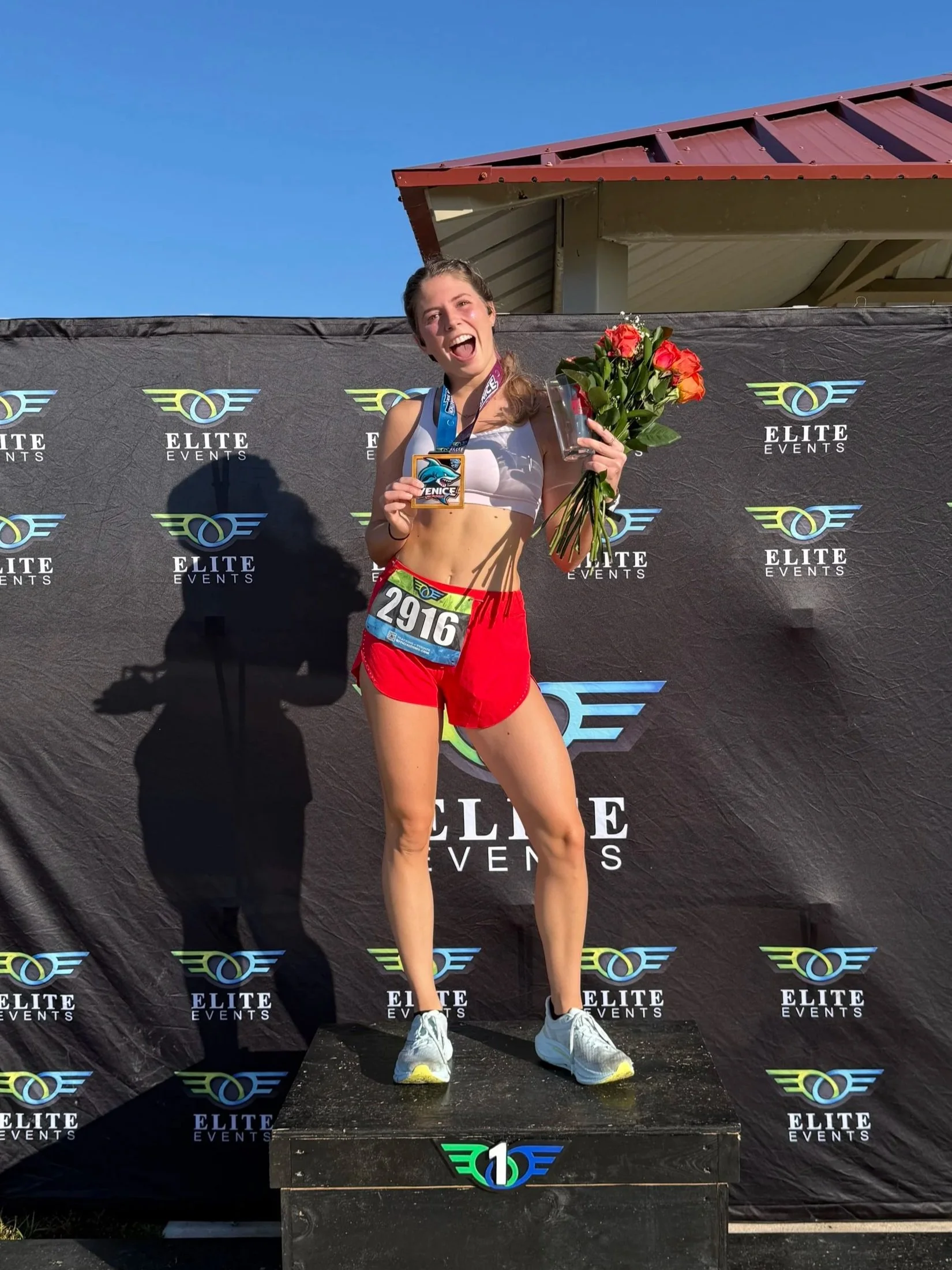 A young woman standing on a winners' podium holding a bouquet of roses and a medal, celebrating her victory at an athletic event, smiling and wearing a sports bra, shorts, and running shoes.