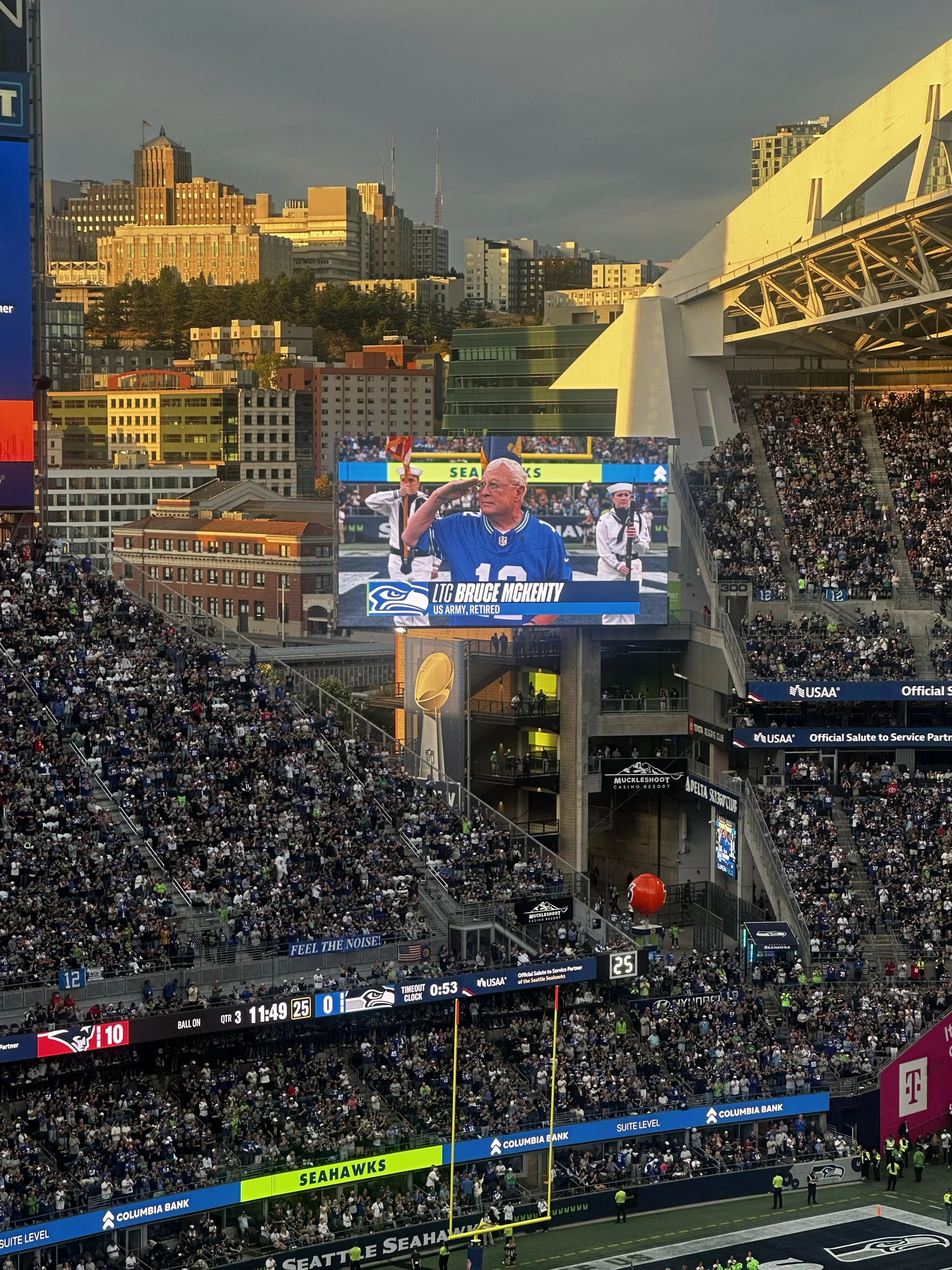 The big screen at Lumen Field shows Bruce McKenty saluting, captioned LTC Bruce McKenty, US Army, Retired, above a full stadium at sunset.