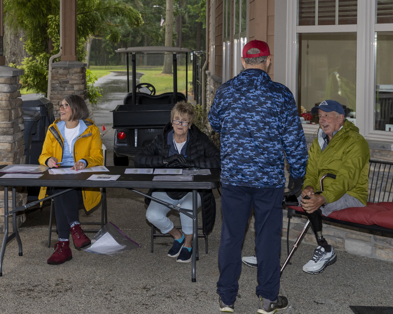 A golfer with a prosthetic leg talks with a man at an outdoor registration table on a rainy day.