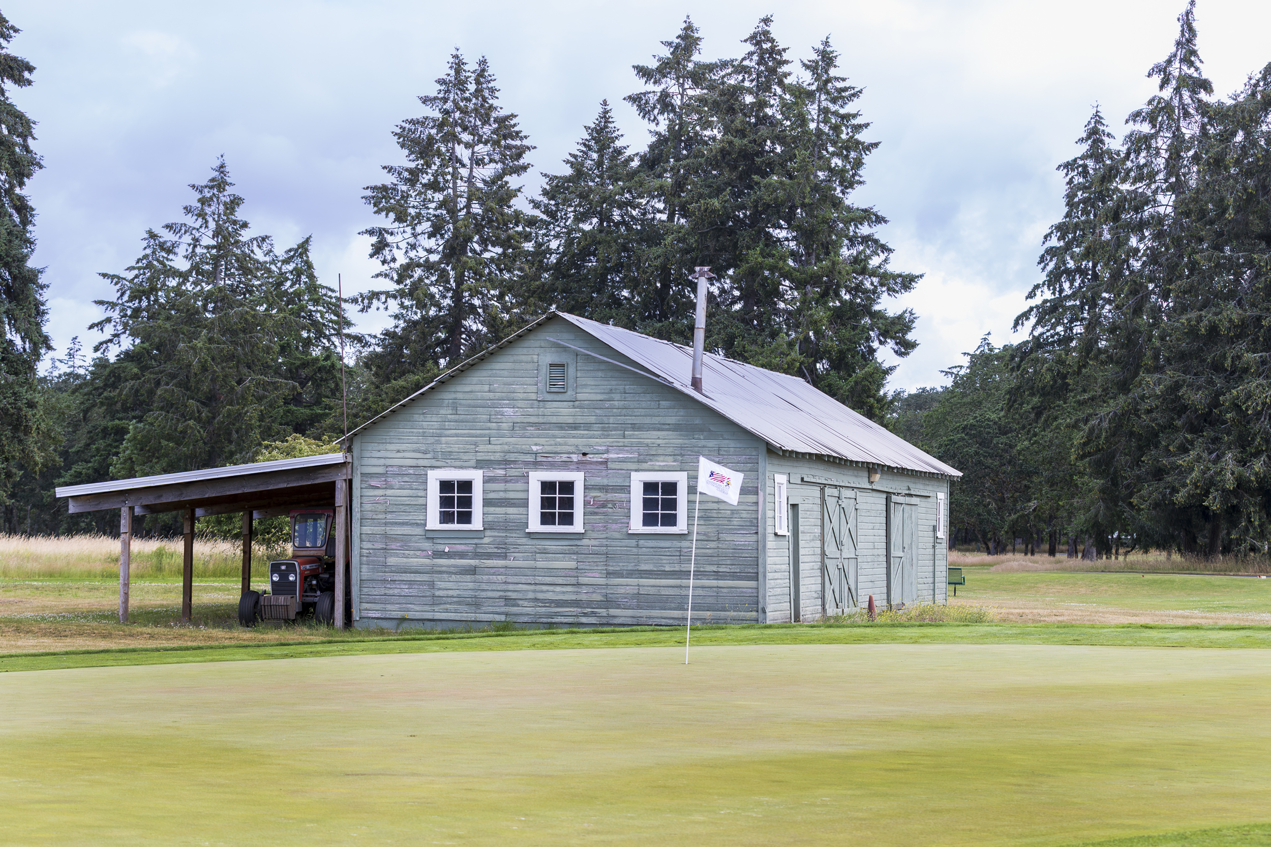 A wooden building with a metal roof beside the green at Hole 16, among tall evergreens.