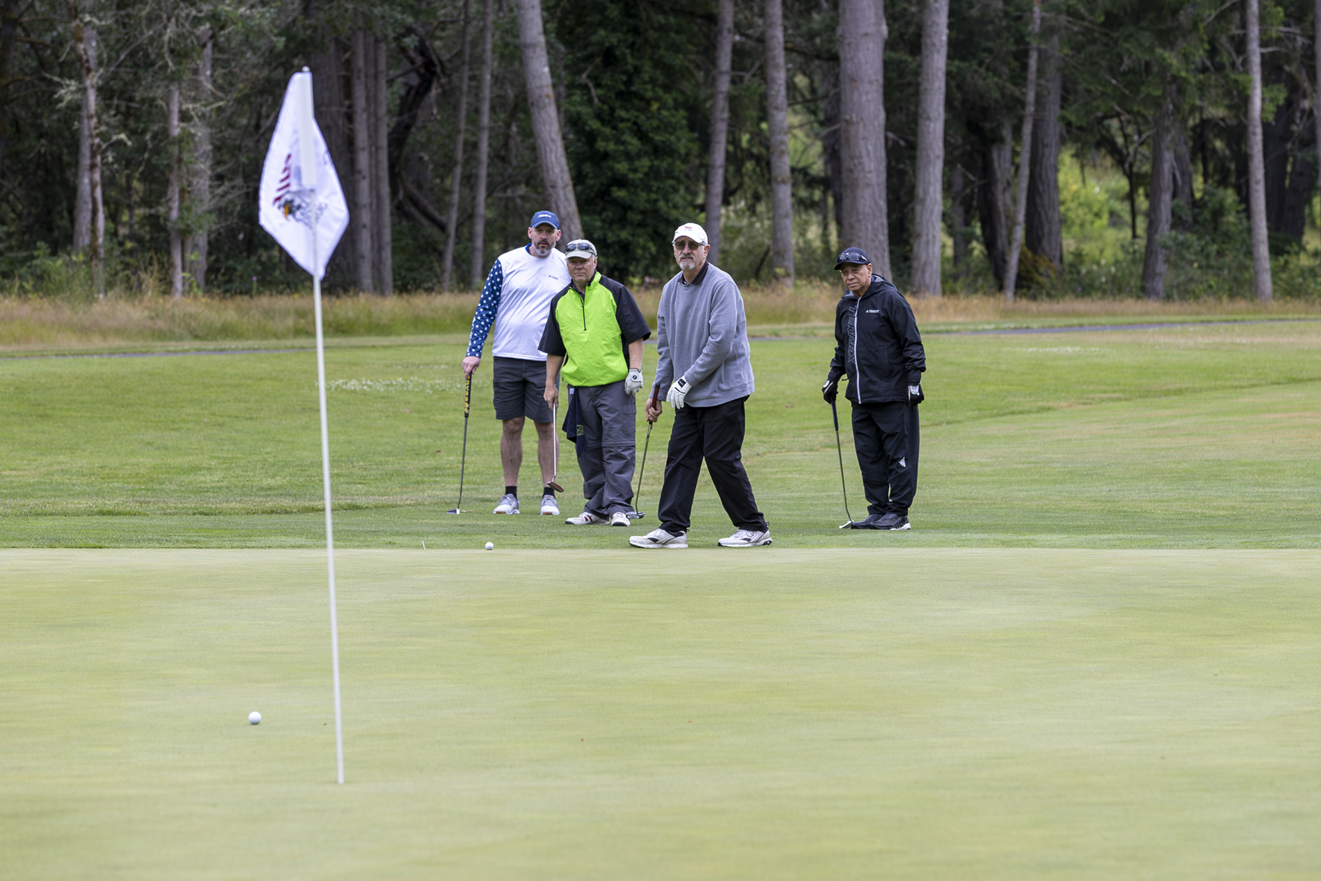Four golfers on a putting green surrounded by evergreen trees.