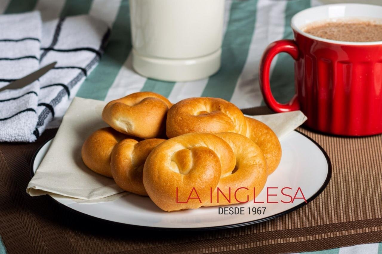 Un plato con cinco panes aliñados tradicionales colombianos tipo Trenza sobre un paño blanco en un plato blanco con borde negro, al fondo una taza roja con bebida caliente, en una mesa con mantel a rayas verdes y blancas.