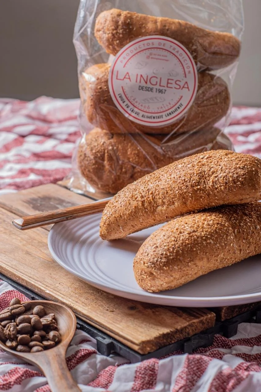 Tres panes de masa horneada cubiertos con costra dorada, colocados en un plato blanco sobre una tabla de madera. En el fondo, un paquete transparente con algunos panes y etiqueta que dice "La Inglesa" desde 1967, en un patrón de tela de cuadros rojos
