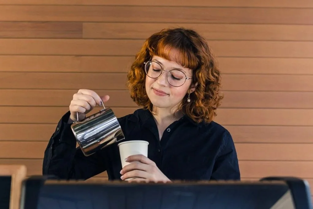 A woman with curly red hair, glasses, and pearl earrings is pouring coffee from a silver pot into a white cup in a cafe or restaurant with wooden wall paneling.
