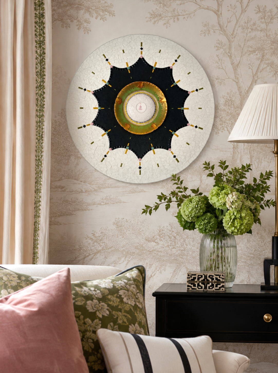 Decorative wall clock with a black star-shaped design on the face, surrounded by a mosaic white circle, hanging on a wall with beige and brown forest scene wallpaper. A vase with green hydrangeas and leafy branches is on a black side table nearby, with a cream-colored sofa and pink and green pillows in the foreground, and a white lampshade on a tall lamp to the right.