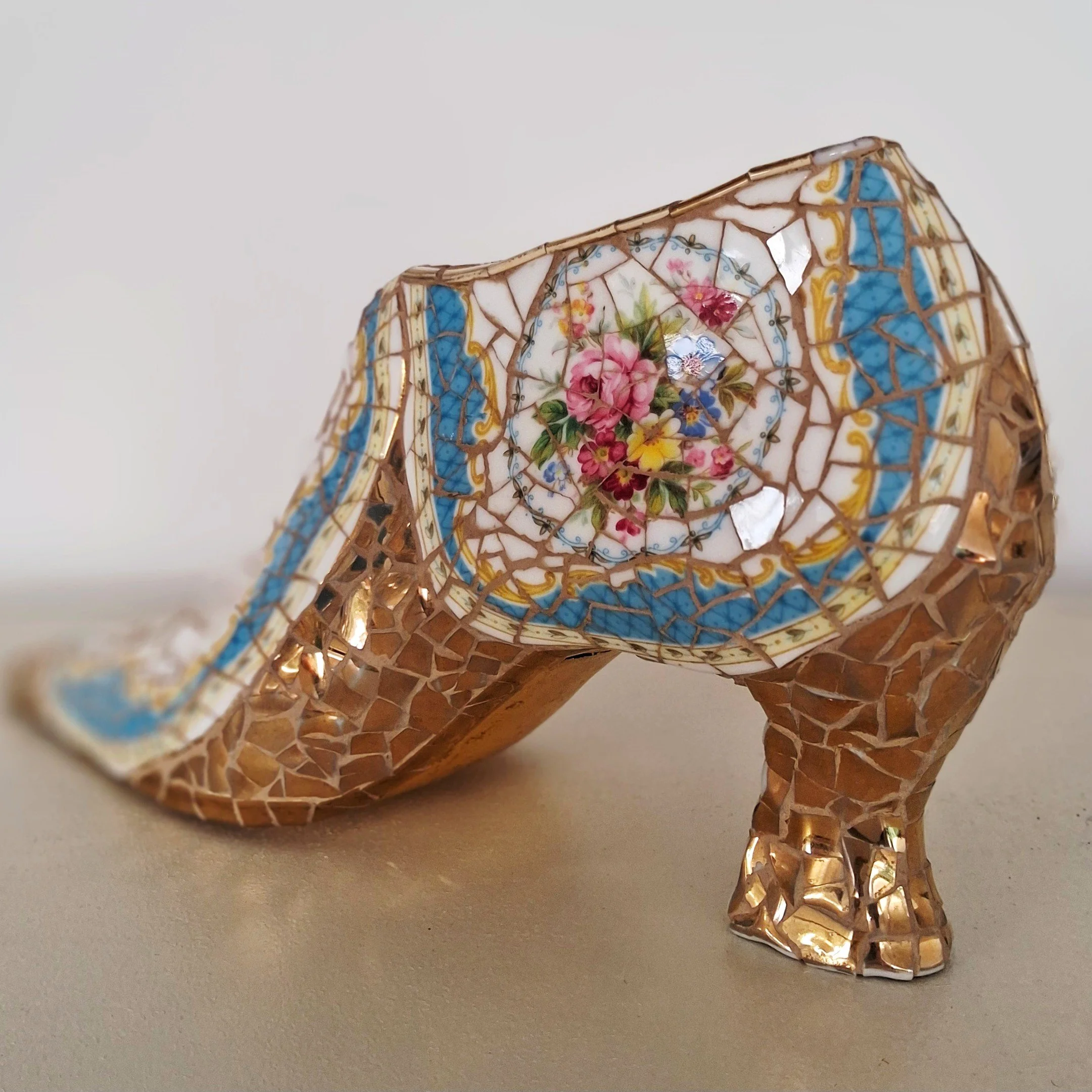 Close-up of a decorative Mosaic Shoe made of vintage china with floral patterns, set against a plain white background.