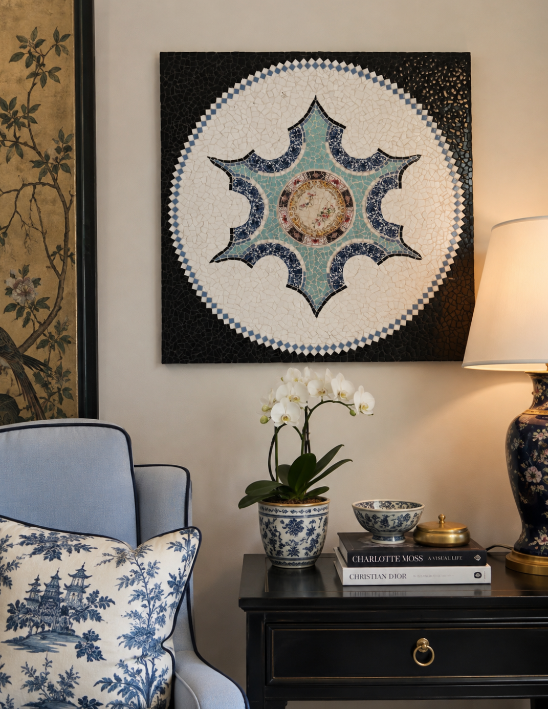 Decorative mosaic wall art above a table with a white orchid in a blue and white pot, a bowl, and books, with a lamp and a pillow featuring blue and white designs nearby.