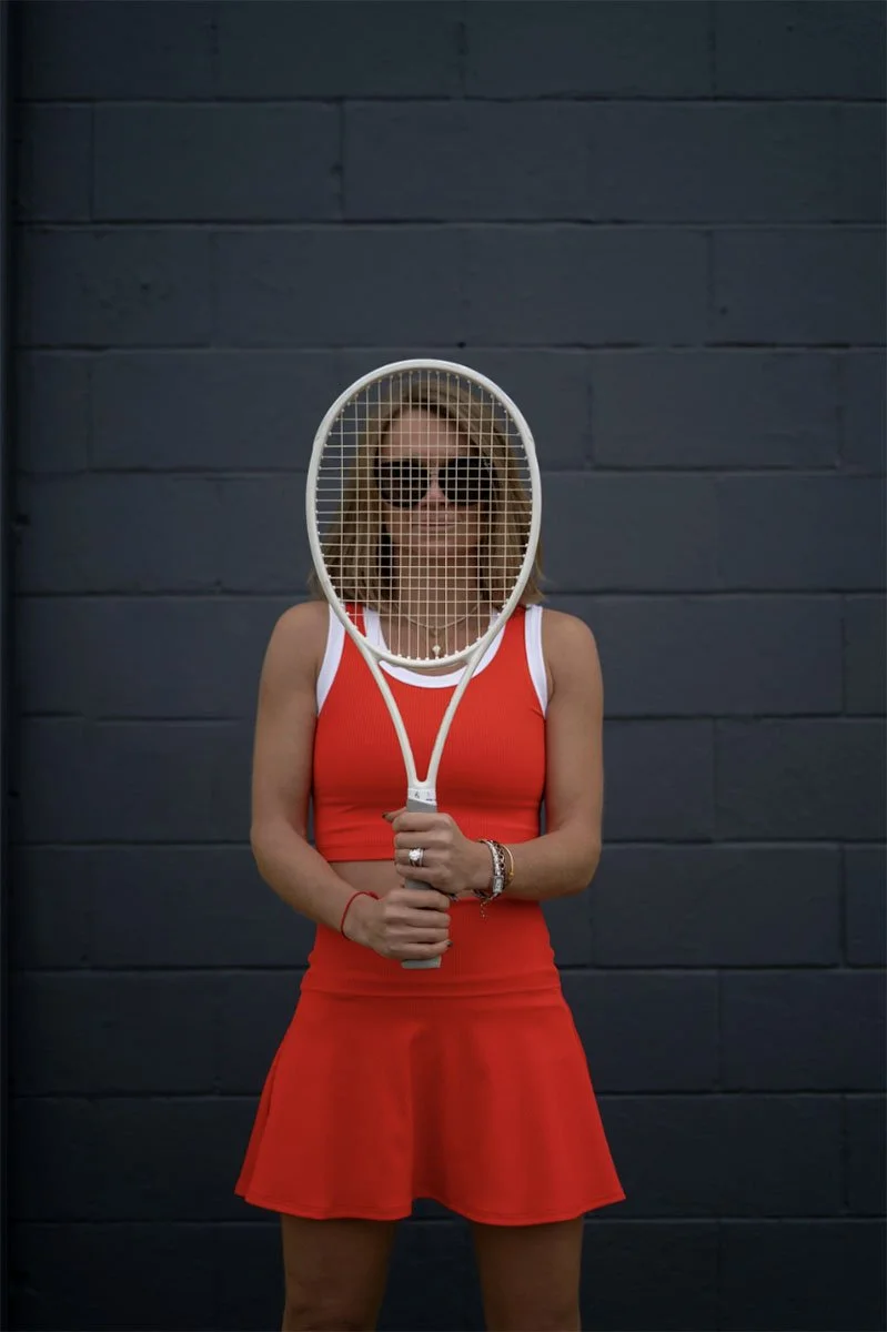 Woman wearing a red sports dress and sunglasses, holding a tennis racket in front of her face, standing against a dark wall.