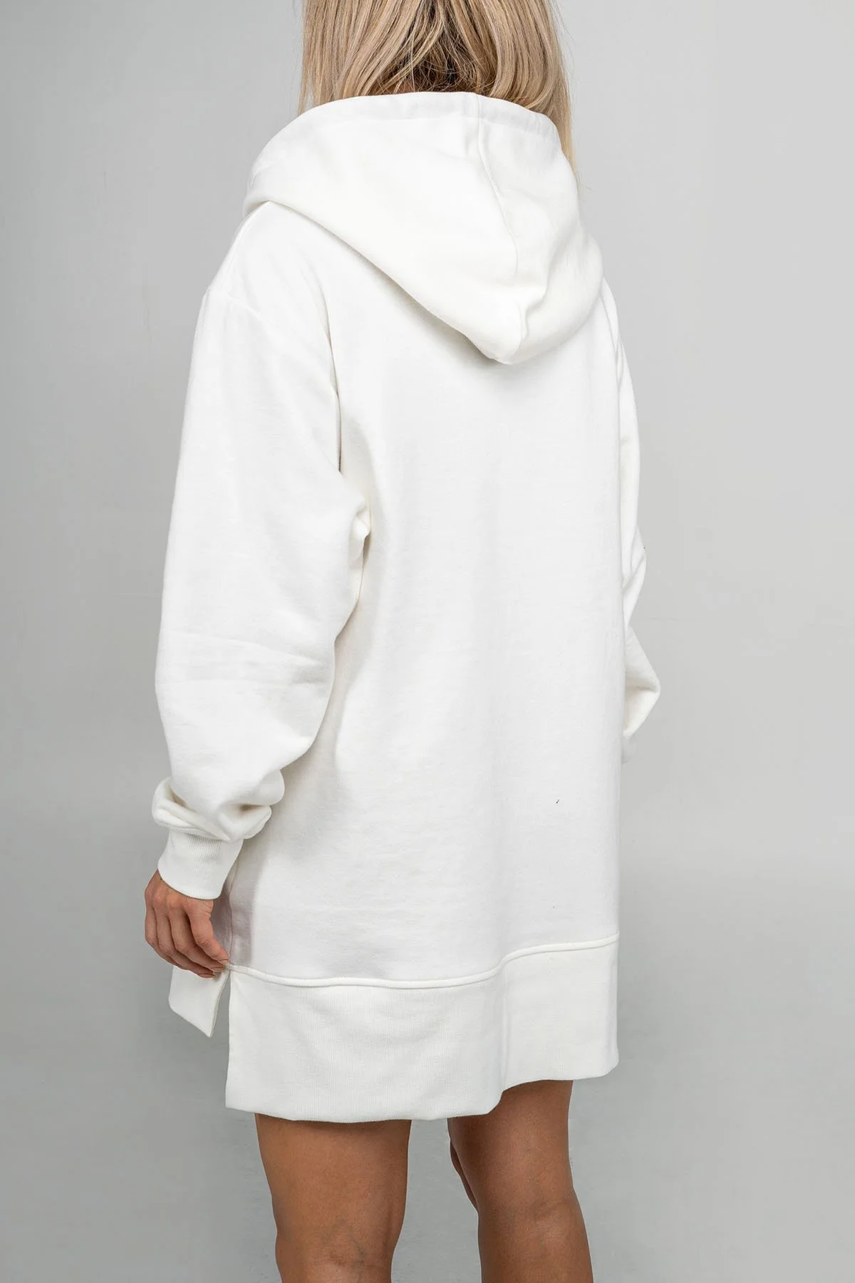 A woman wearing a white hoodie and matching white skirt, standing against a plain light gray background, seen from the back.