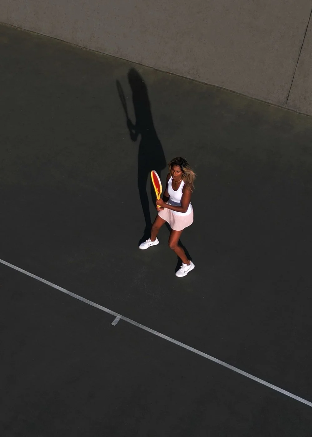 A woman holding a tennis racquet on a tennis court during daytime, casting a long shadow.