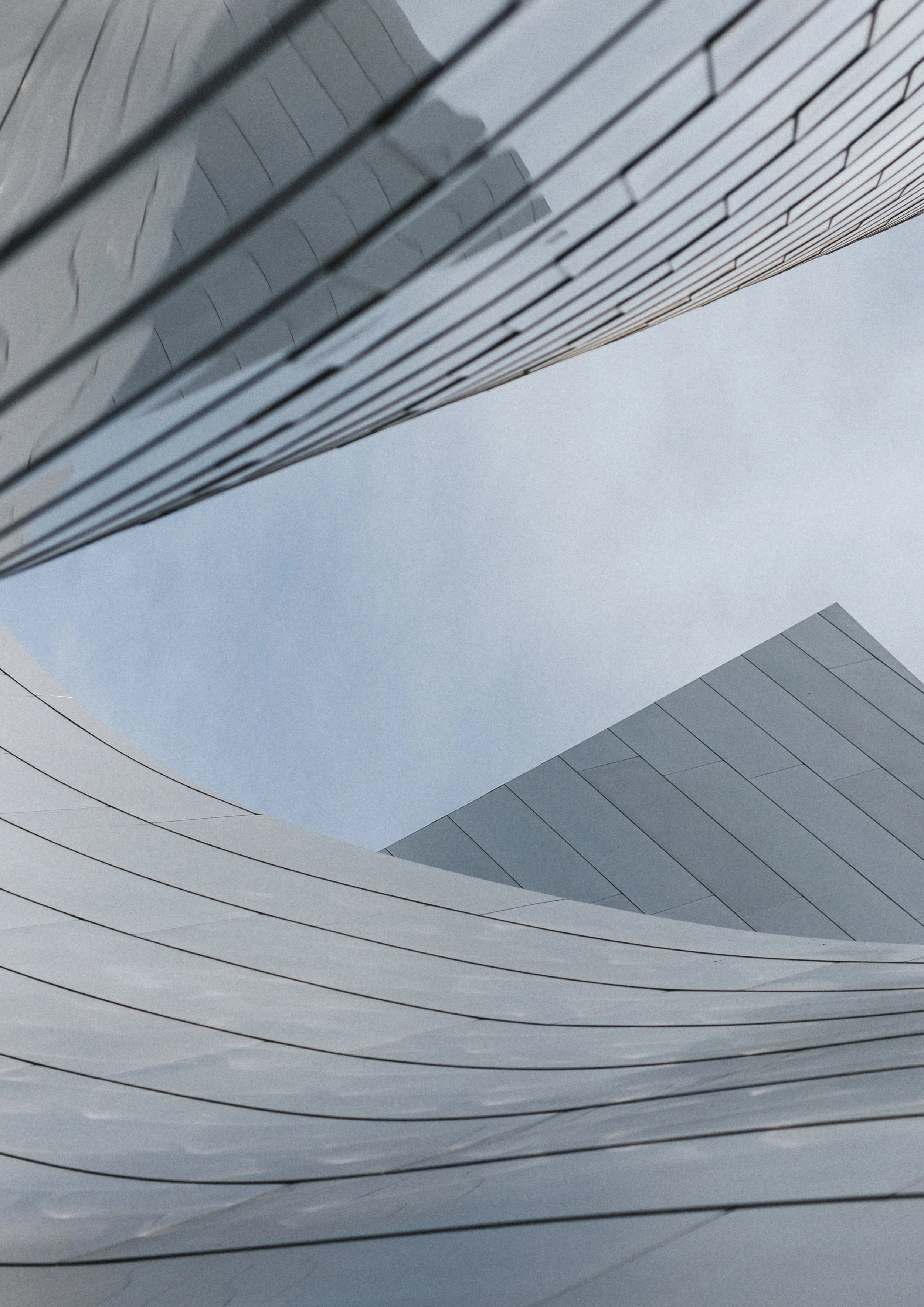 Close-up of the curved exterior of a modern building made of white and light grey panels with thin black lines, viewed from below against a cloudy sky.