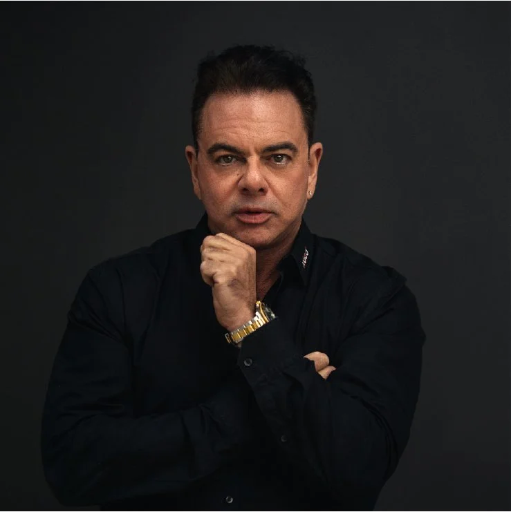 A man with dark hair, wearing a black shirt, gold watch, and earring, posing with his hand on his chin against a dark background.