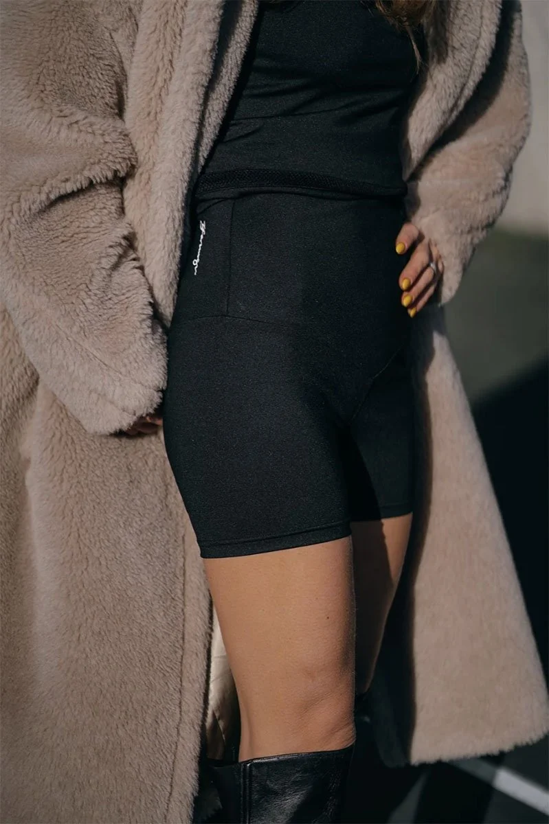 Person wearing a beige fuzzy coat, black top, black shorts, and black knee-high boots.