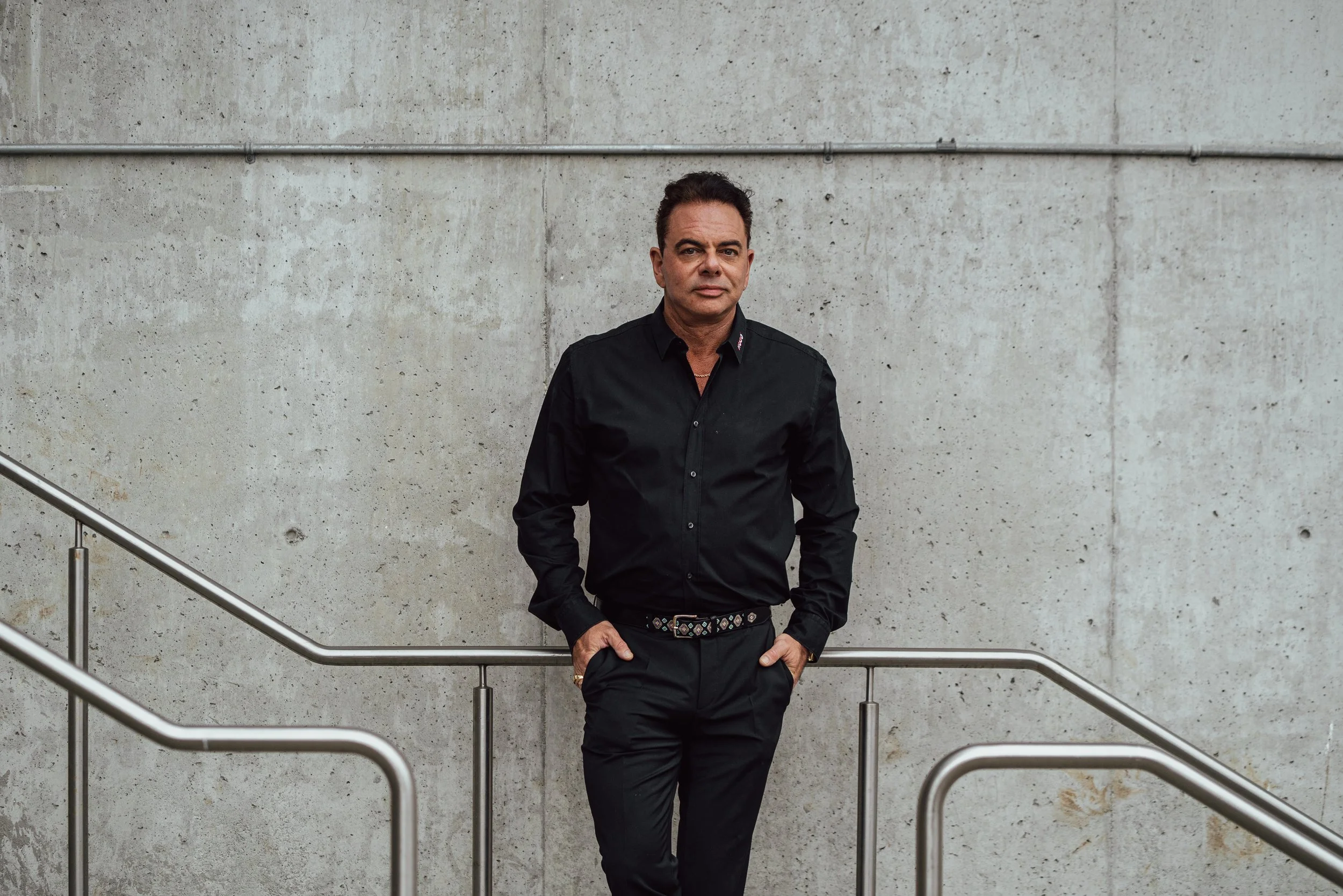 A man in black clothing standing next to a metal railing, with a plain concrete wall in the background.
