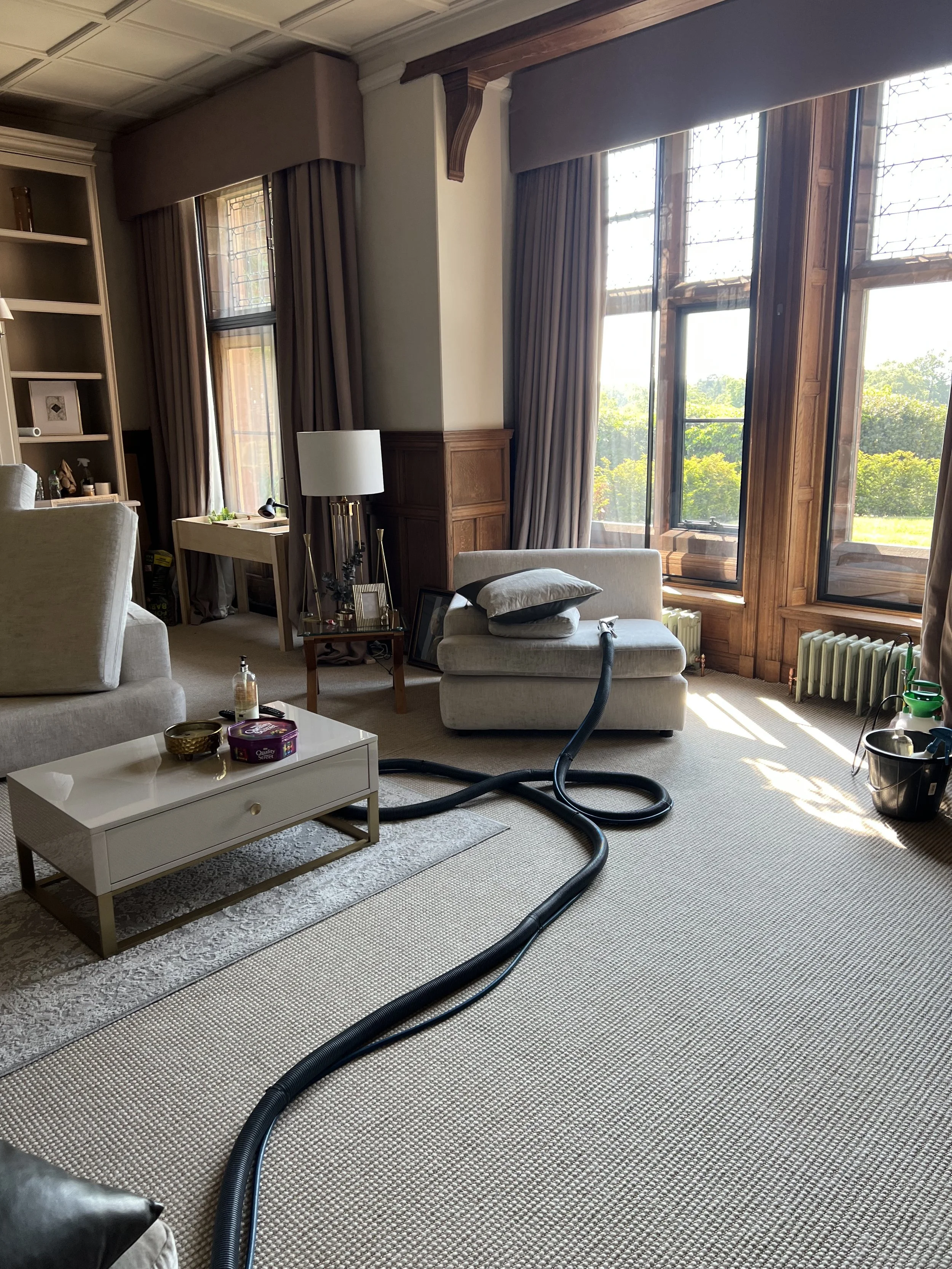 Living room with large windows, beige carpet, beige sofa, white coffee table, floor lamp, bookshelf, and vacuum hose on the floor.