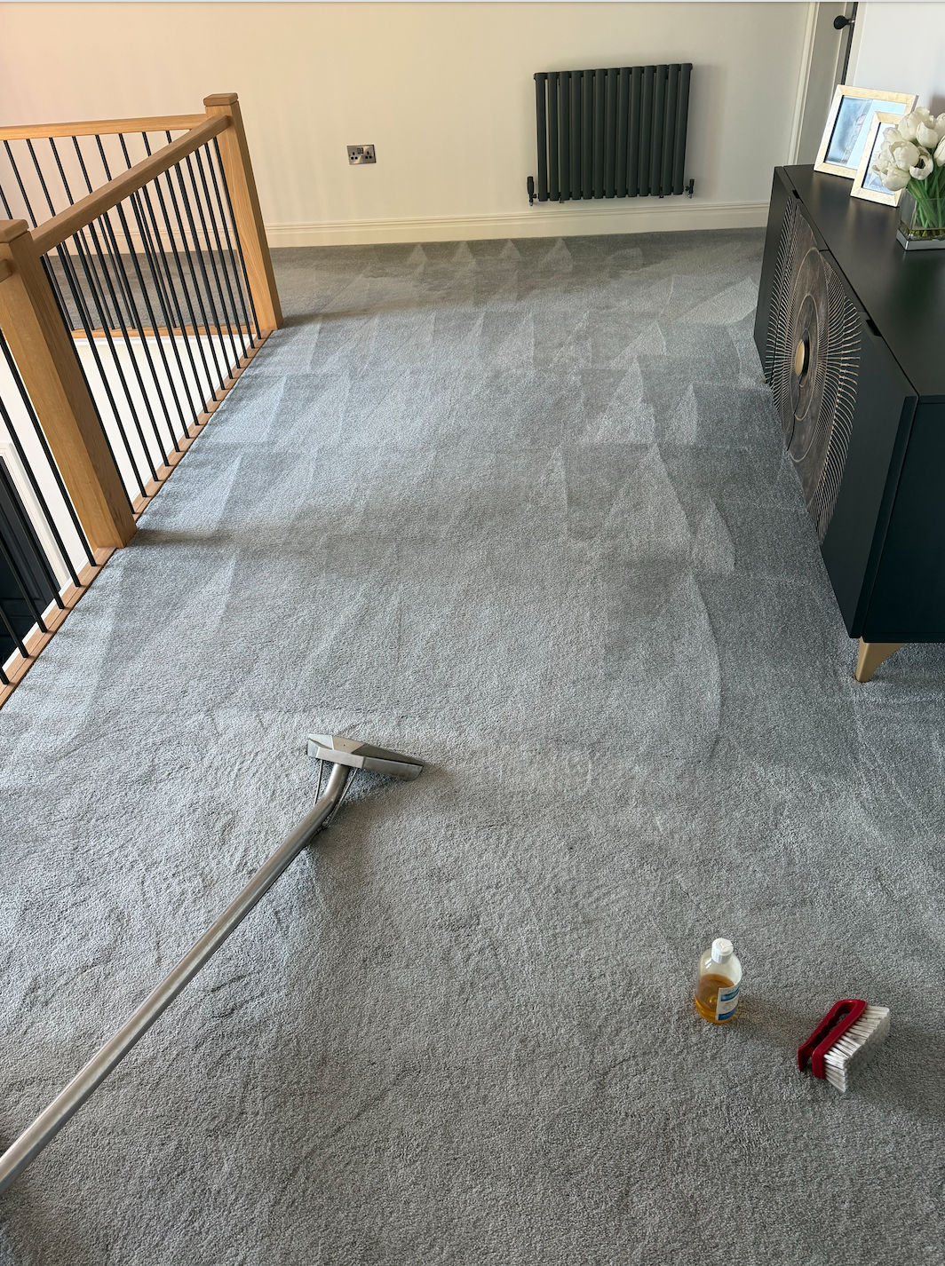 A light gray carpet being vacuumed with a gray vacuum cleaner. A bottle of cleaning solution and a small wire brush are on the carpet. The room has a wooden staircase, a black radiator, a black sideboard, and decorative items like framed photos and white flowers.