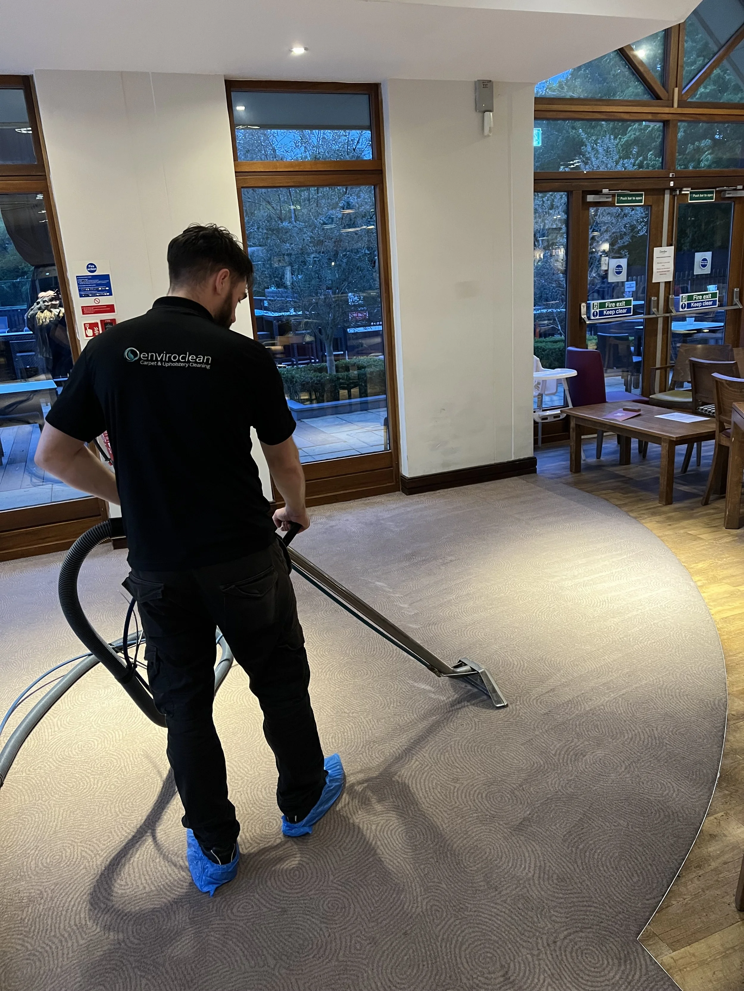 A man wearing blue shoe covers and black pants vacuuming a beige carpet in a well-lit room with large windows and wooden frames, and a view of trees outside.