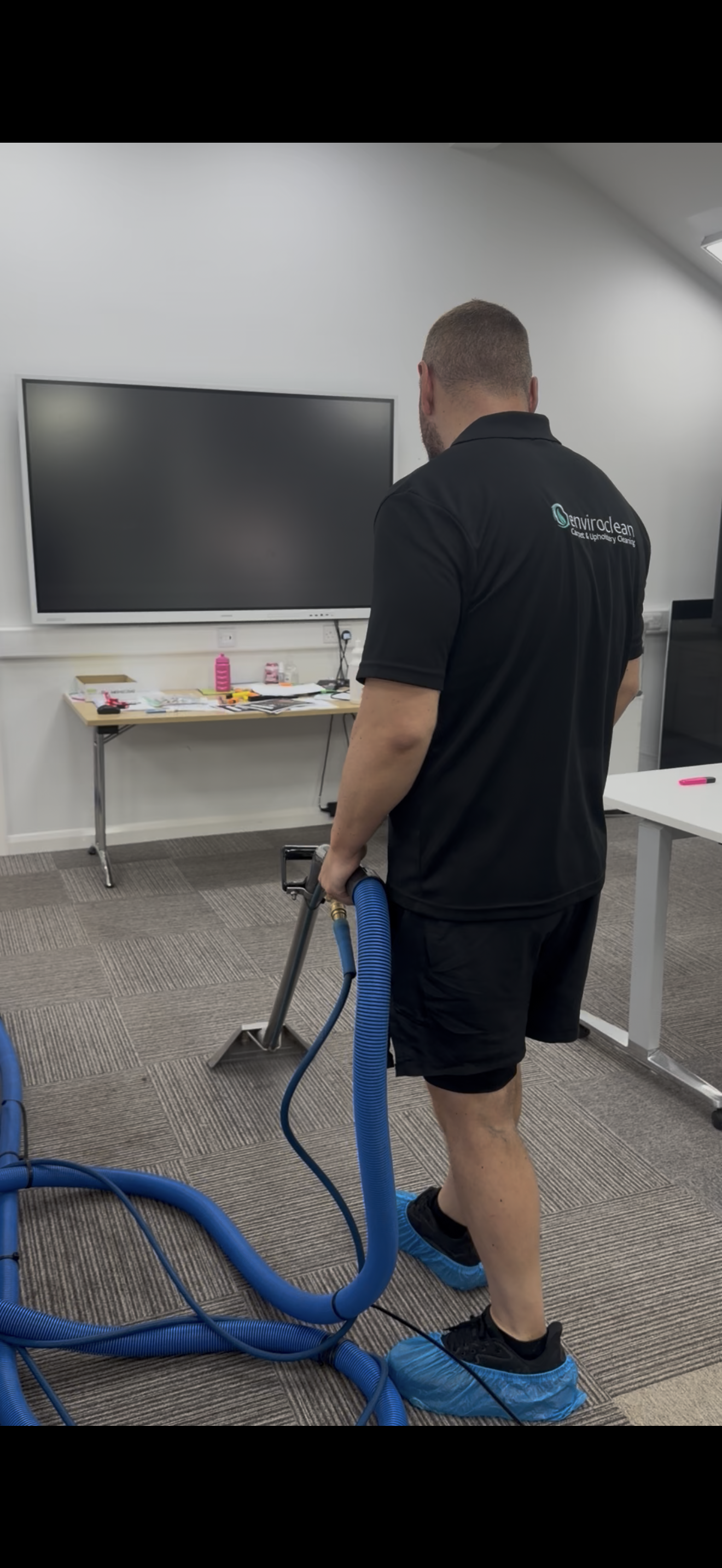 A man is standing in an office or conference room, wearing black clothing and disposable shoe covers, holding a blue wet vacuum hose. In the background, there is a large flat-screen TV mounted on the wall and a table with cleaning supplies.