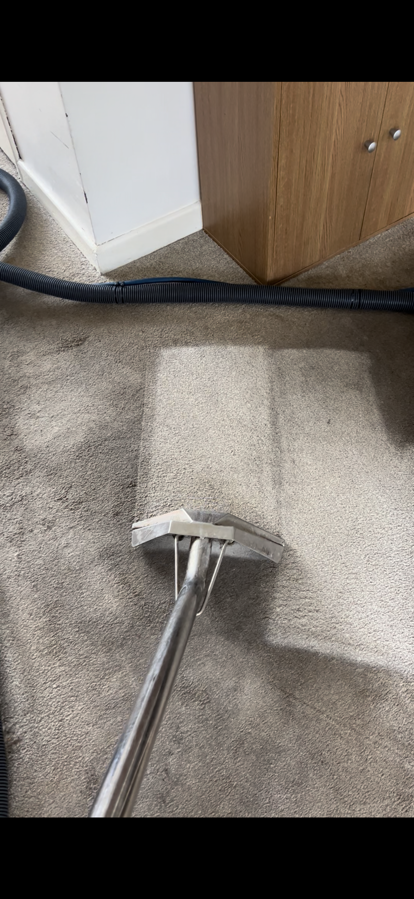 Close-up of a carpet cleaning wand on beige carpet with a floor vent, a black vacuum hose, and a wooden cabinet in the background.
