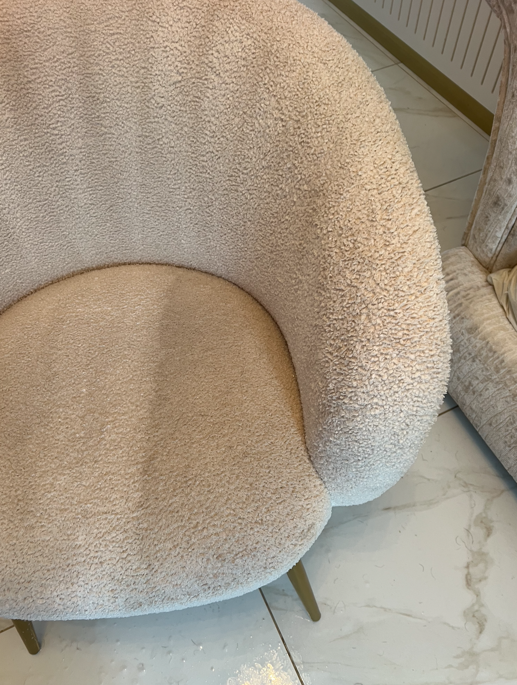 Close-up of a beige, textured upholstered chair with round seating and a curved backrest, on a marble floor.