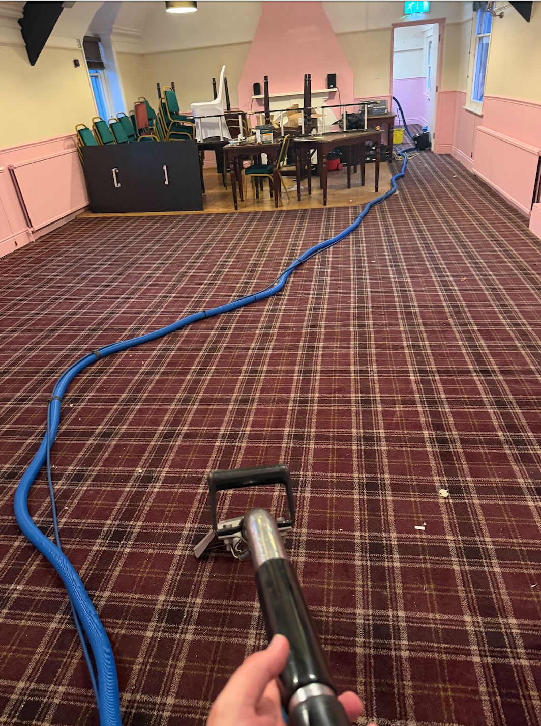 Person cleaning a carpeted room with a steam machine connected to a long blue hose inside a decorated room with chairs stacked along the wall, tables, and a pink fireplace.