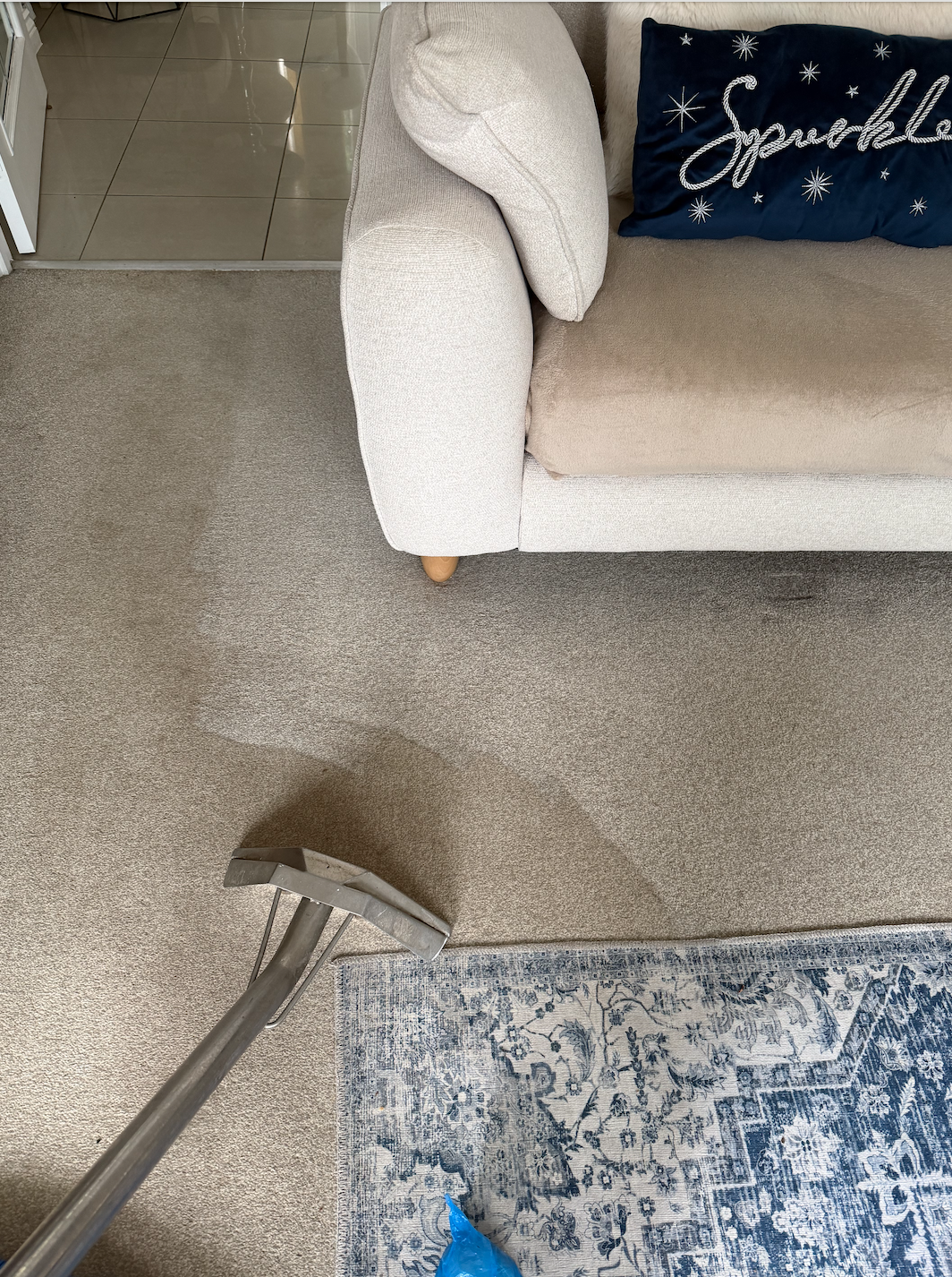 A person vacuuming beige carpet in a living room with a beige armchair, a beige sofa with a navy blue pillow that says 'Sparkle' and has star designs, and a patterned blue and white rug. The vacuum cleaner attachment is held in hand, and the room leads into a tiled area visible through an open doorway.