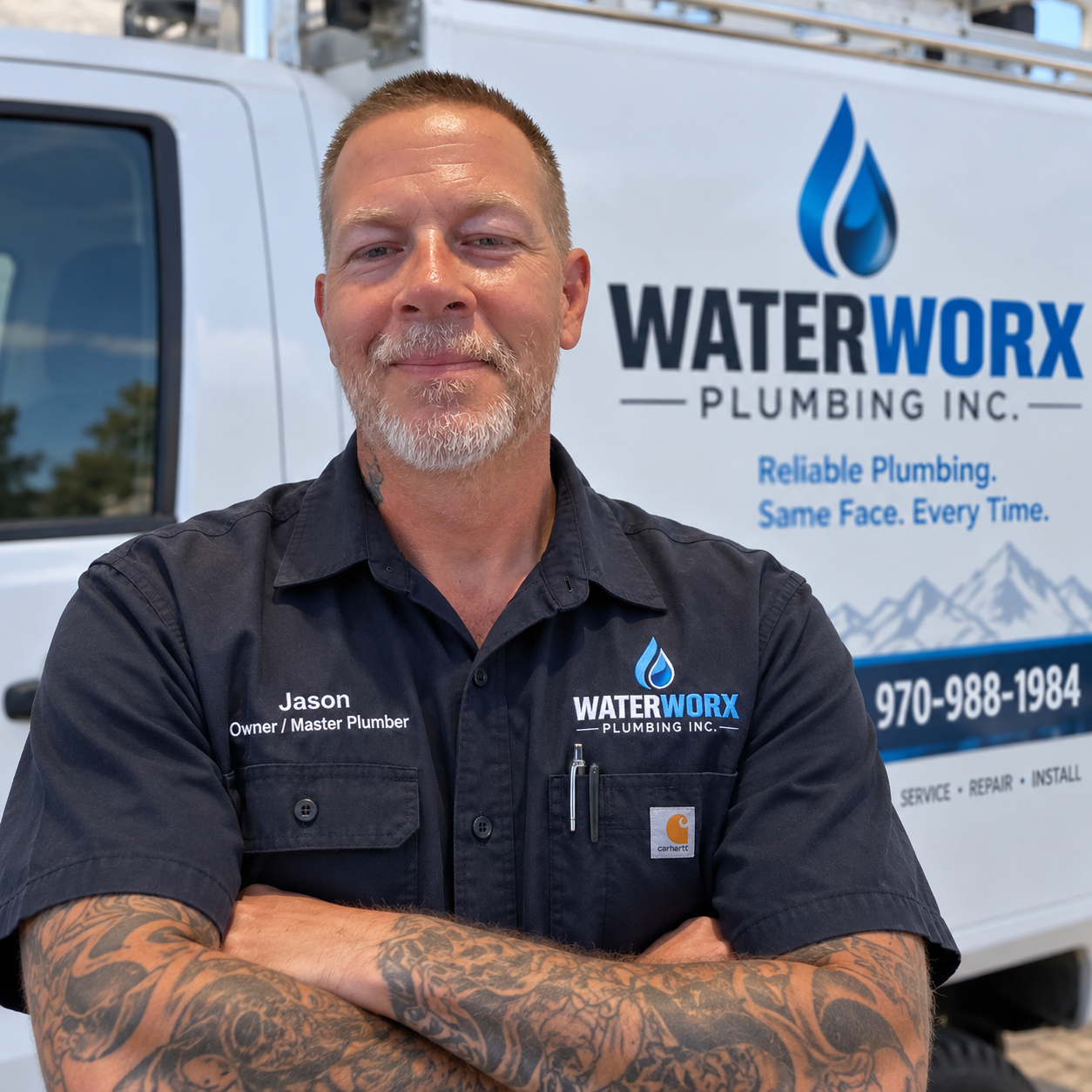 A man with a beard and tattoos crossing his arms, wearing a black shirt with the logo and name tag of WaterWorx Plumbing, standing in front of a water delivery truck with the company's branding and contact information.