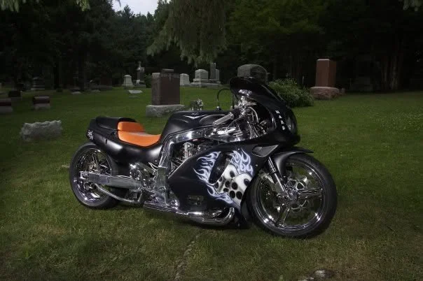Motorcycle with black and white flame design on fairings and an orange seat parked on grass near headstones in a cemetery.