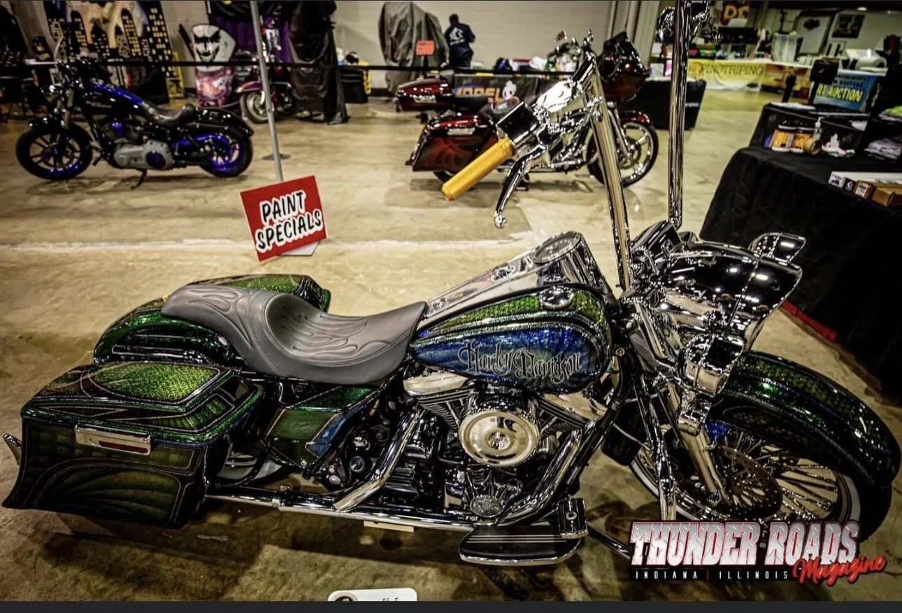 A custom Harley Davidson motorcycle with green, blue, and black paint, chrome accents, and a comfortable seat, displayed at an indoor motorcycle show with other bikes and booths in the background.