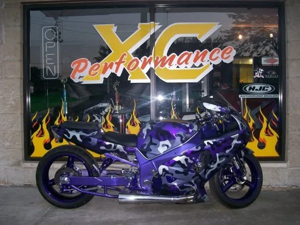 A purple and black camouflage motorcycle parked outside a performance shop with a large window display featuring flames and the words 'XC Performance' and various stickers.