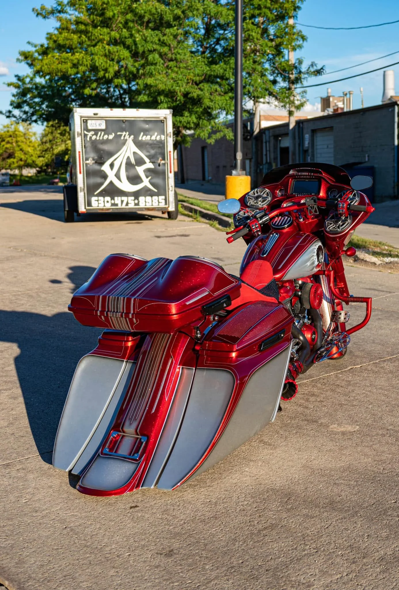 A red motorcycle with matching red and silver fairings parked on a concrete driveway. In the background, there is a black trailer with white writing and logo, green trees, and buildings.