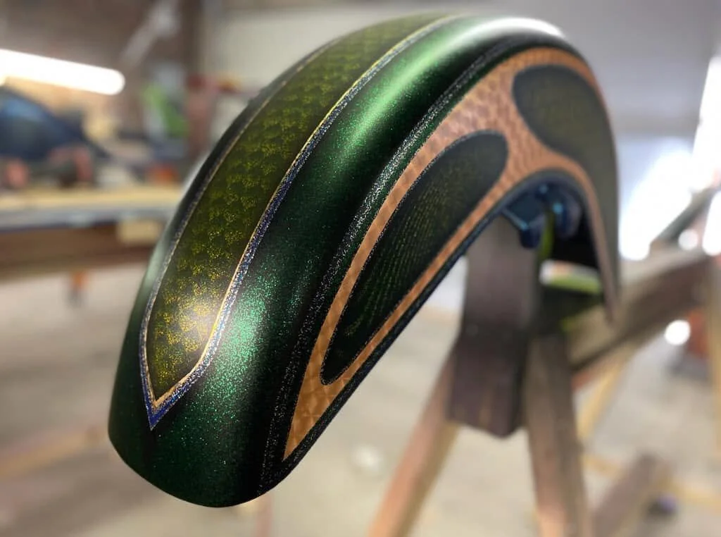 Close-up of a custom-painted musical instrument part, with green, gold, and black detailing, mounted on a wooden stand inside a workshop.