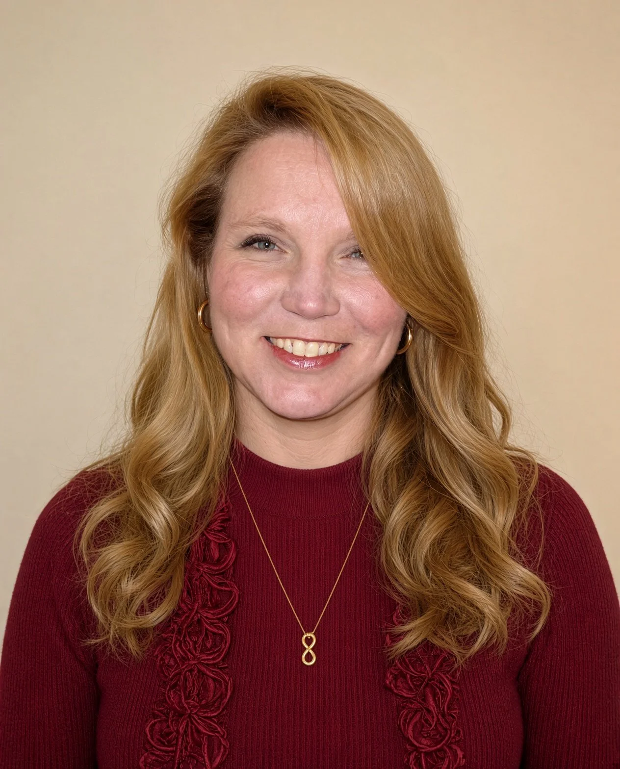 A woman with long wavy red hair, wearing a burgundy sweater with ruffled detail and gold hoop earrings, smiling against a plain beige background.