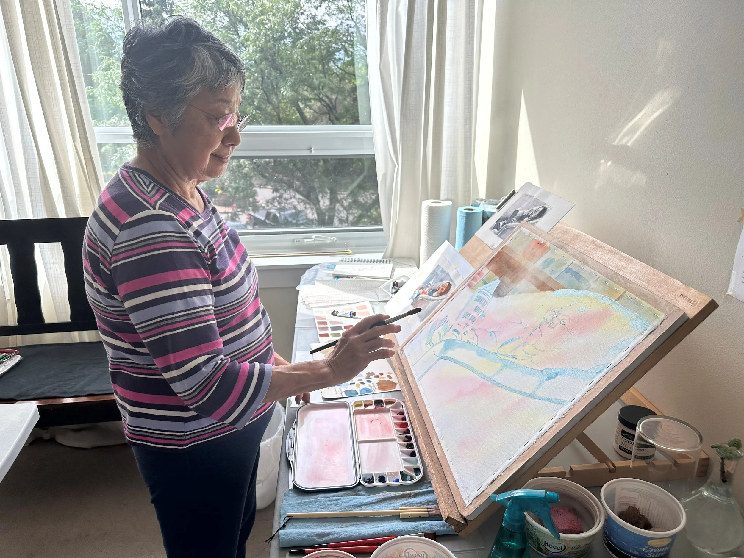 An elderly woman with short gray hair and pink glasses, wearing a striped shirt, working on a watercolor painting at an art studio. She is standing in front of an easel, holding a paintbrush, with various art supplies and watercolor paints on the table.