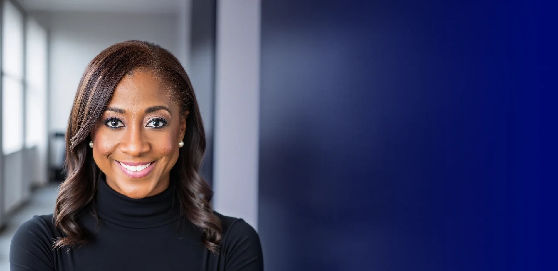 Close-up portrait of Rosilyn "Roz" Houston smiling - with brown hair, wearing pearl earrings and a black turtleneck, in an office setting.with natural light and blue wall background.