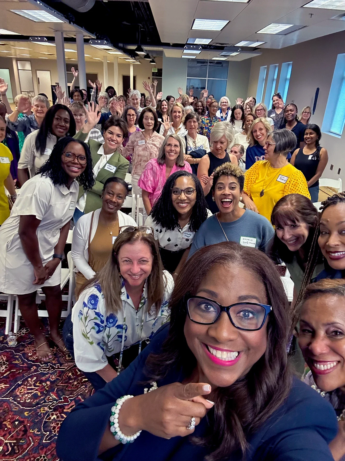Roz with Women for Alabama, smiling and waving at a conference or gathering in a brightly lit room with windows and patterned carpet.