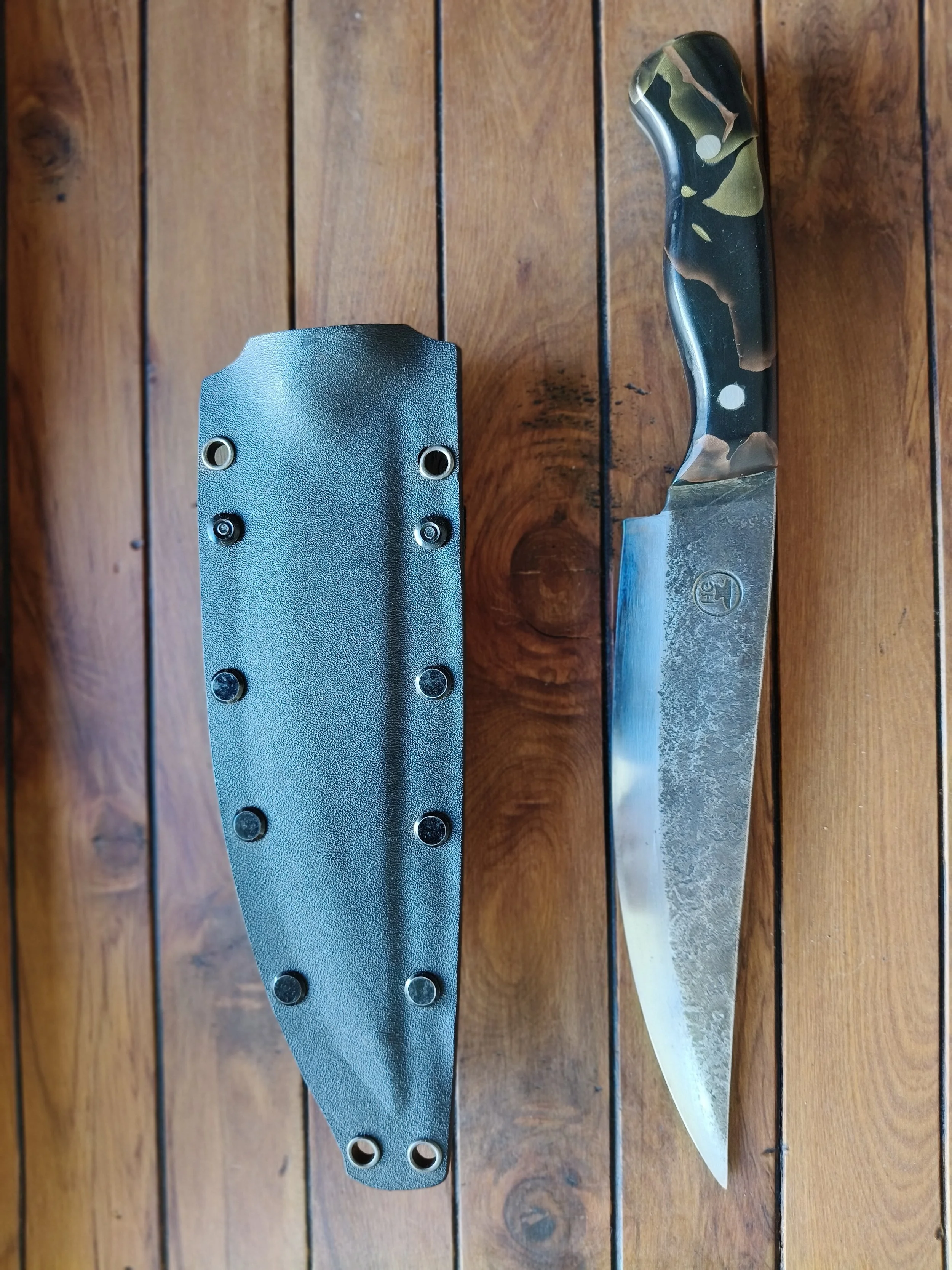 A large kitchen knife with a black handle and a textured metal blade placed on a wooden surface, beside a sharpening stone with black rubber base and metal surface.
