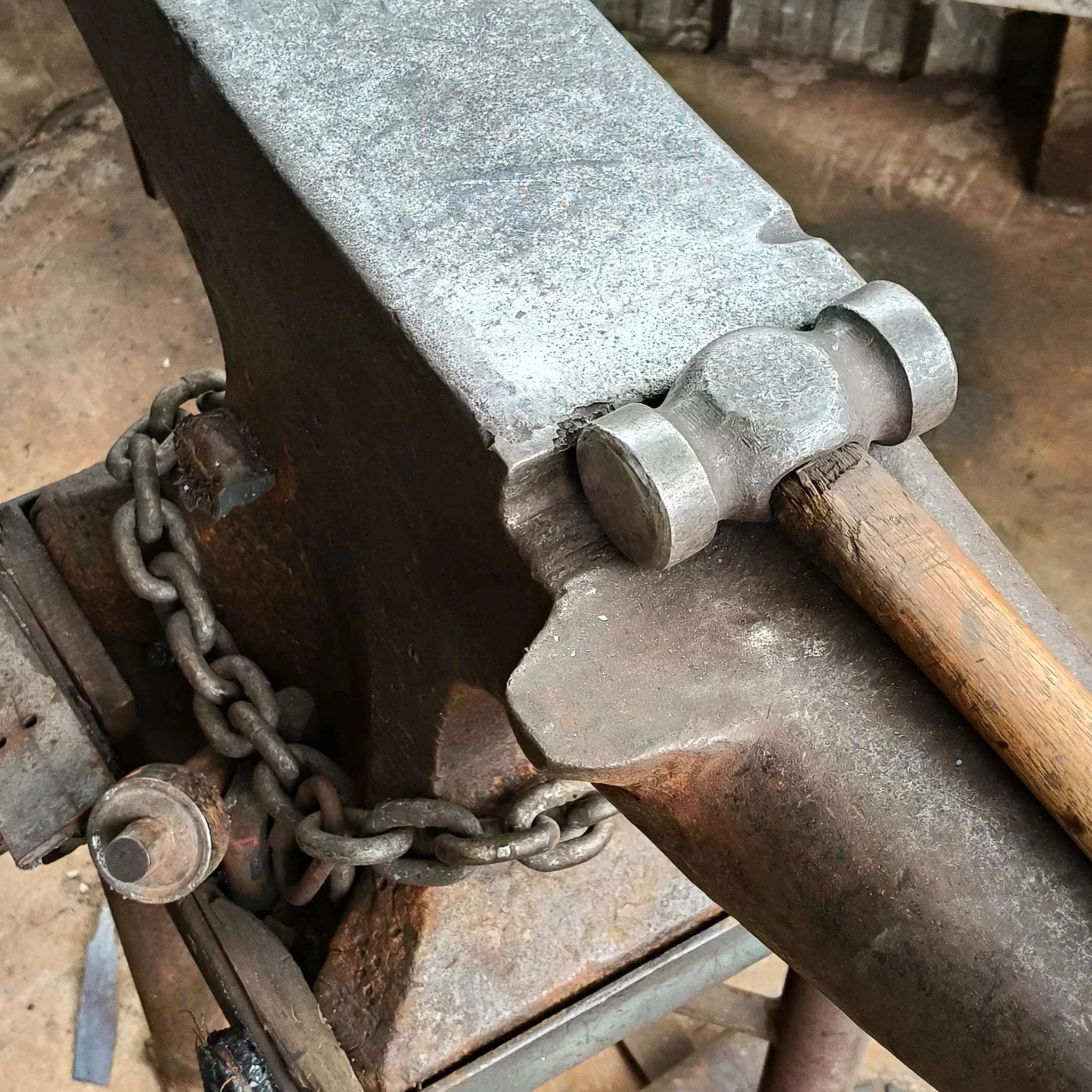 Close-up of a metal forging hammering on an anvil, with a chain and wood handles visible, in a blacksmith workshop.