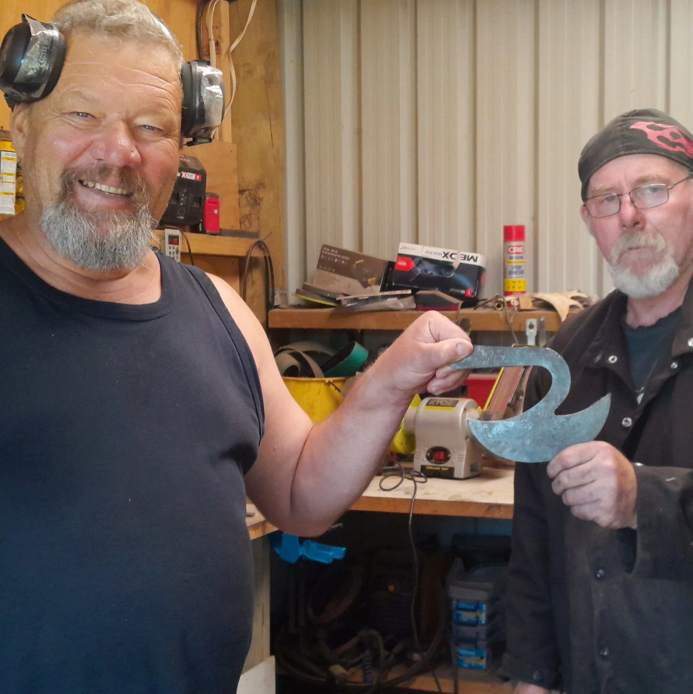Two older men in a workshop, one with a beard and wearing a tank top and ear protection, holding a power tool, and the other with glasses and a bandana, looking at the camera.