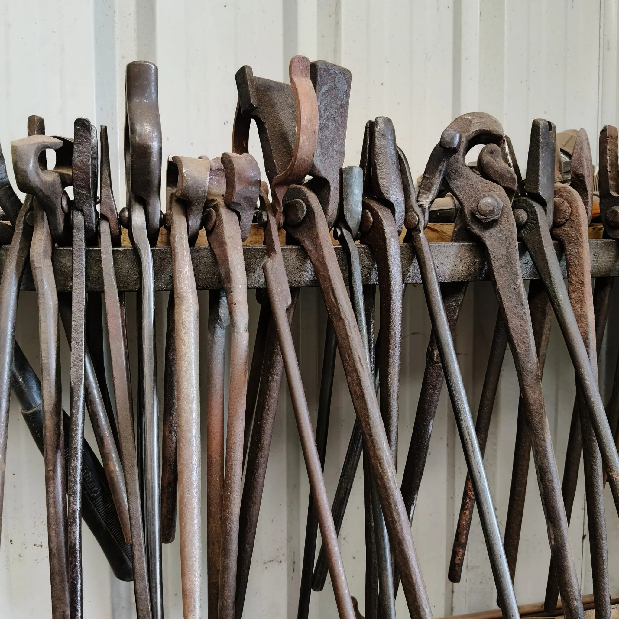 A row of rusty and worn metal hand tools, including clamps, laid out on a wooden surface against a light-colored, vertical-paneled wall.