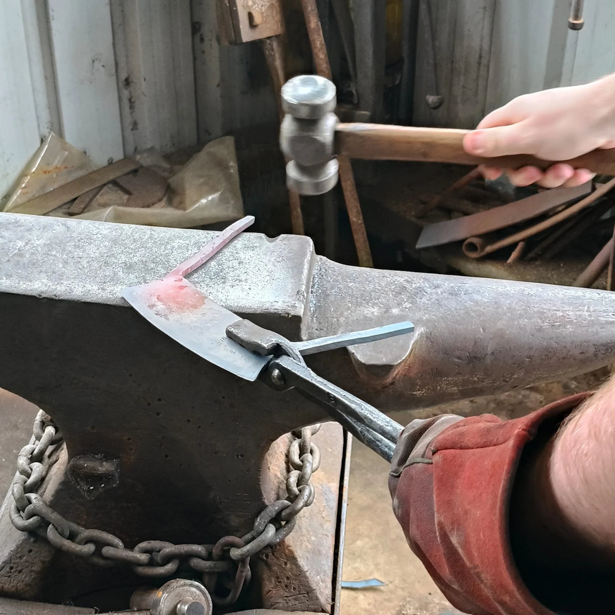 A person wearing red work trousers using a hammer to tighten or loosen a bolt on a machinery component surrounded by chains in an industrial or workshop setting.