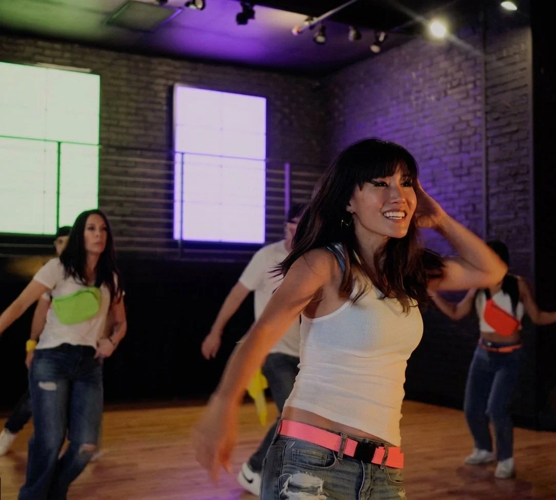 A group of women dancing in a dimly lit studio with brick walls and colorful LED screens on the wall.