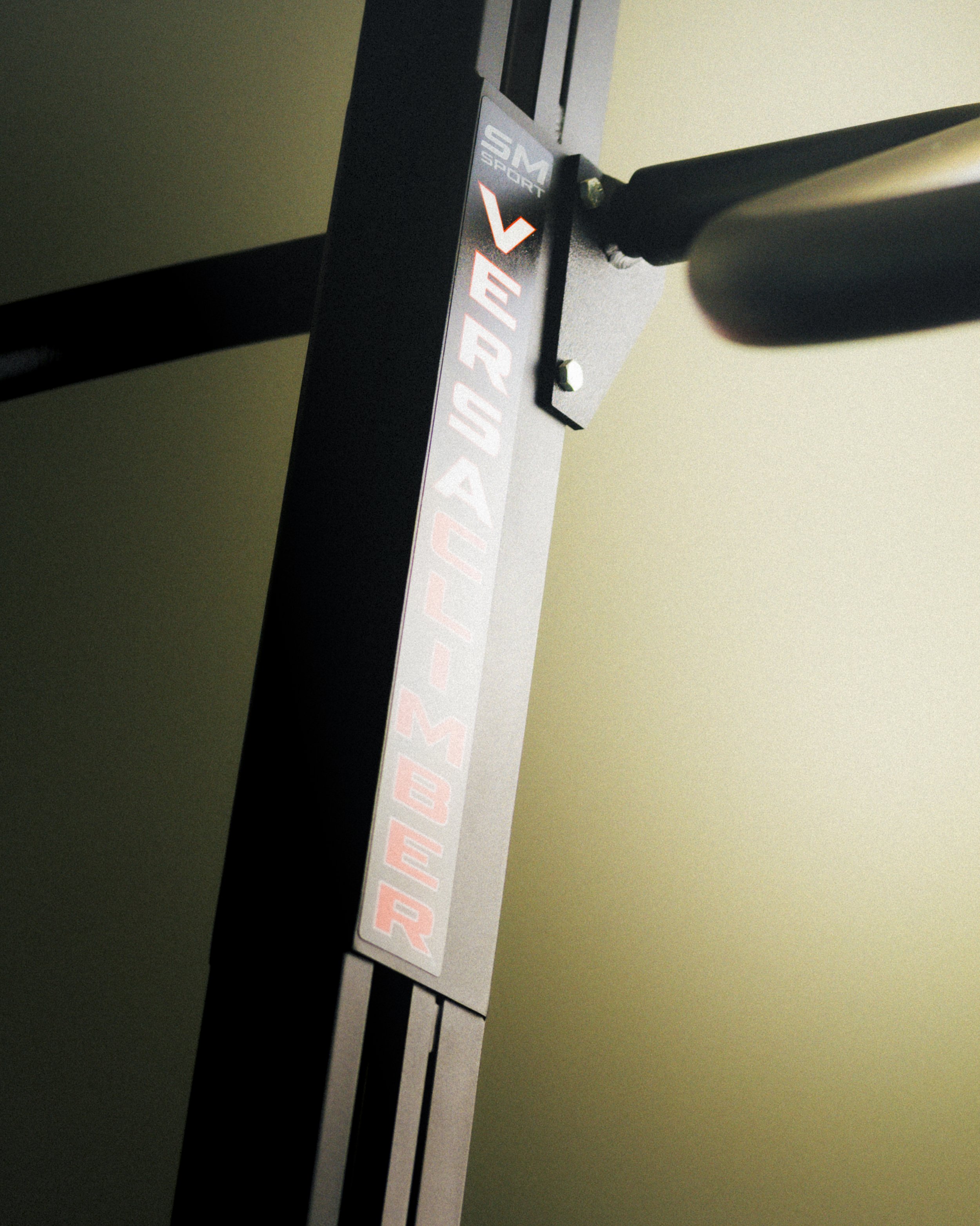 Close-up of a crafting or electronic device with a label that reads "THIS SIDE UP" and a small logo reading "SM SPORT." The device is mounted on a wall with visible screws and has a black handle or arm attached to the right side.