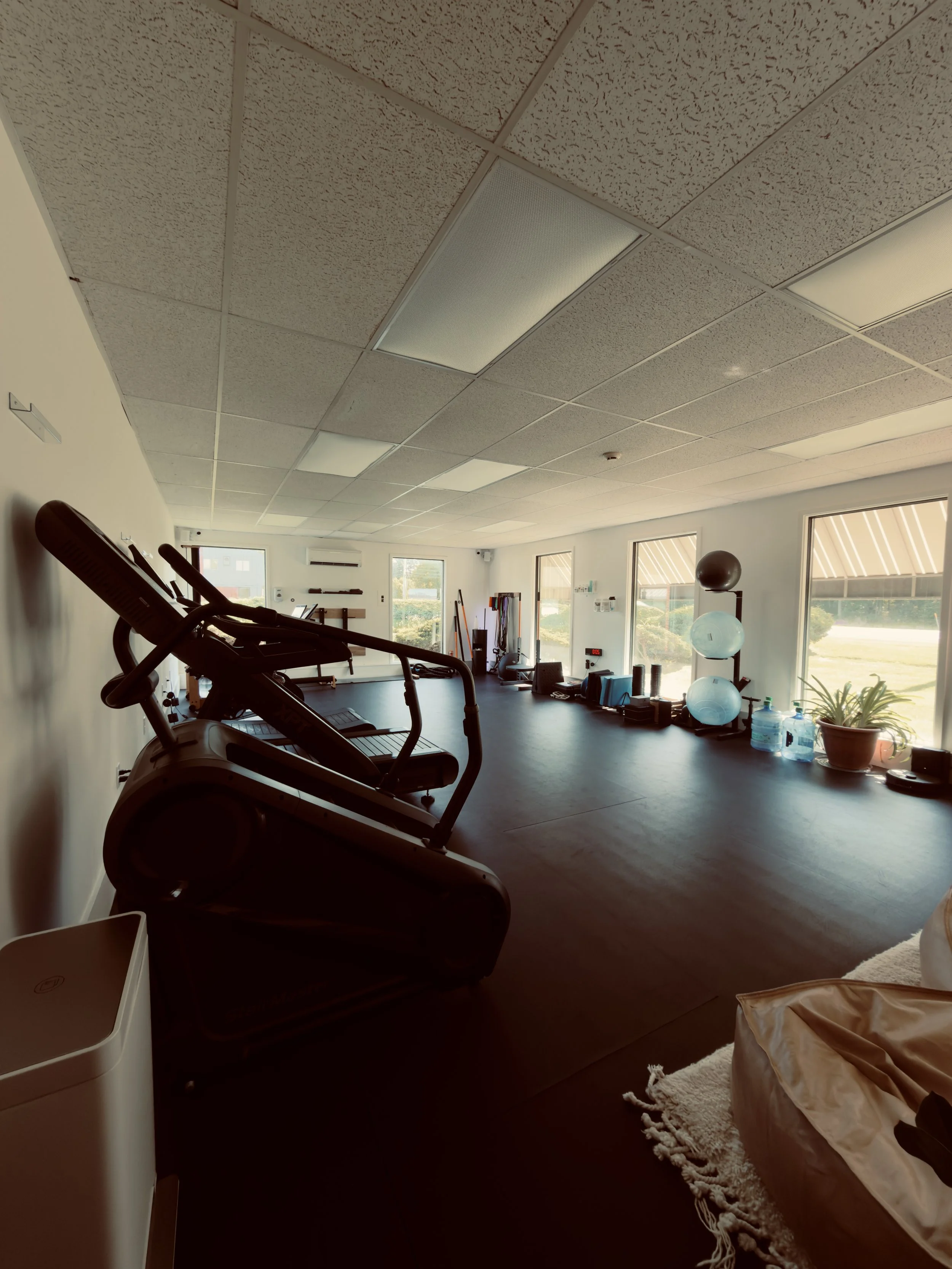 A fitness room with exercise equipment including a treadmill and a stability ball, large windows allowing natural light, and various exercise accessories and water bottles.