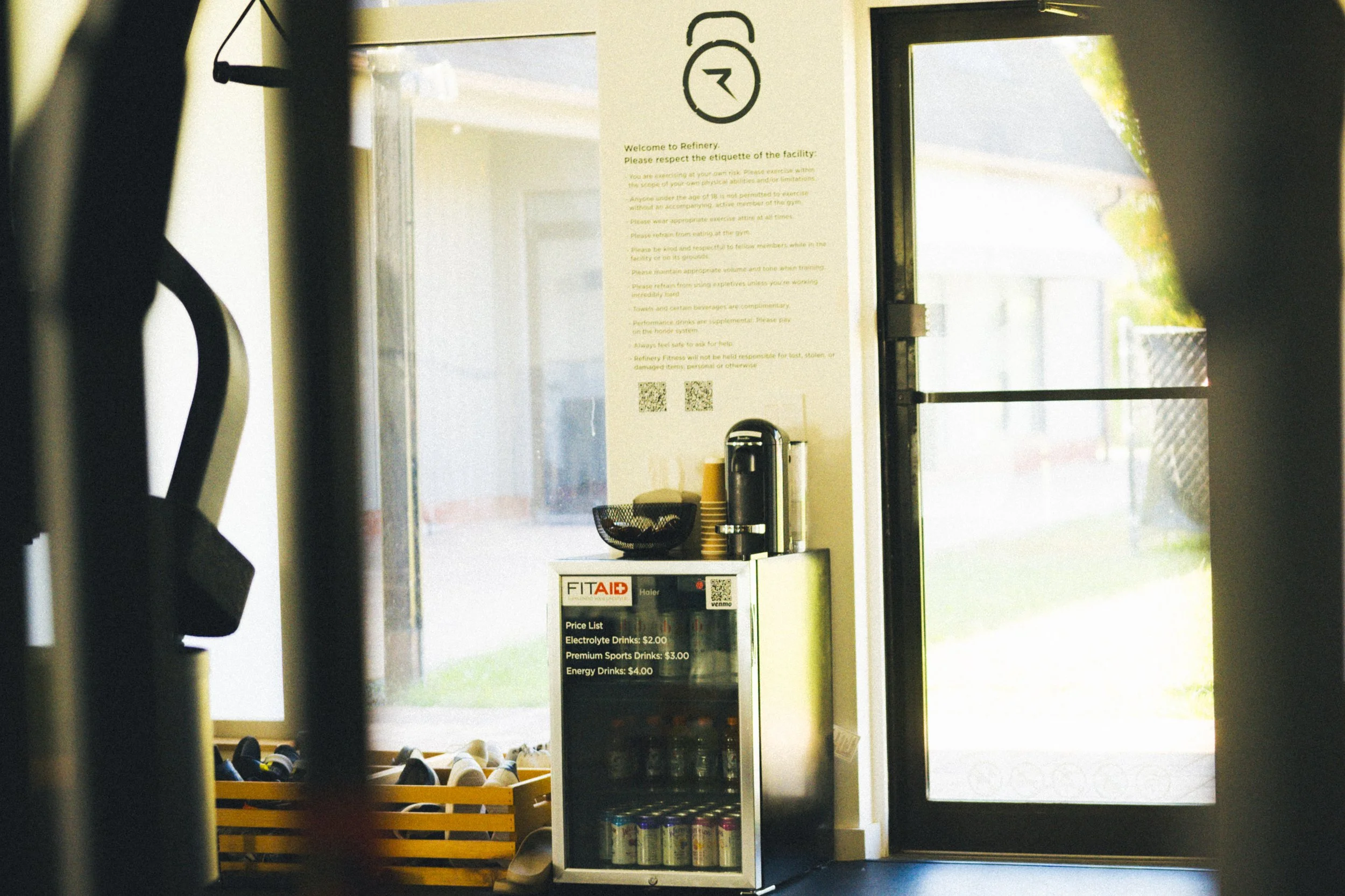 View of the entrance area of a fitness or gym facility with a menu displaying prices for electrolyte, premium sports, and energy drinks, seen through a glass door, with sunlight streaming outside.