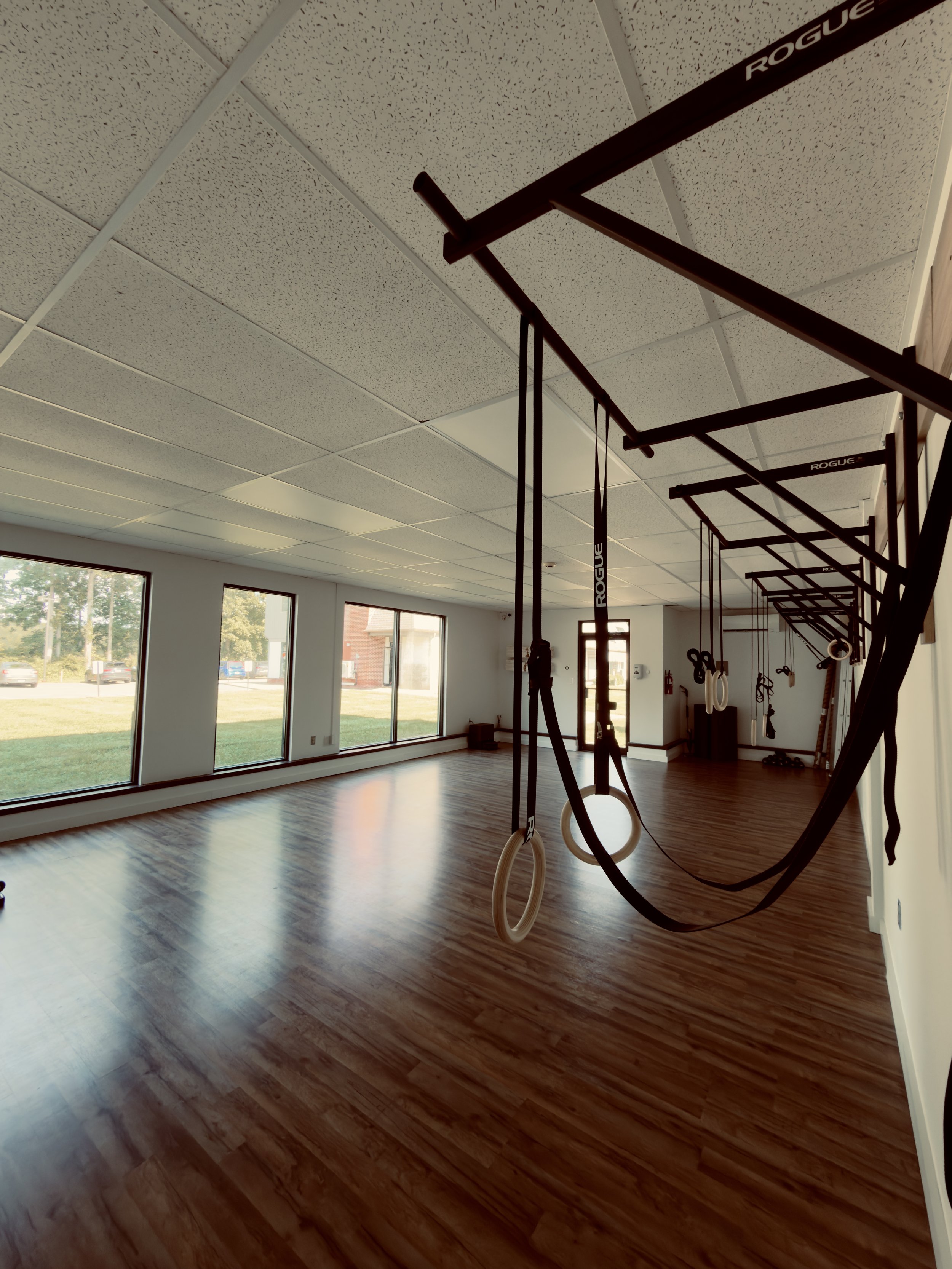 Empty gym with large windows, wooden floor, and gymnastic rings hanging from a red frame.
