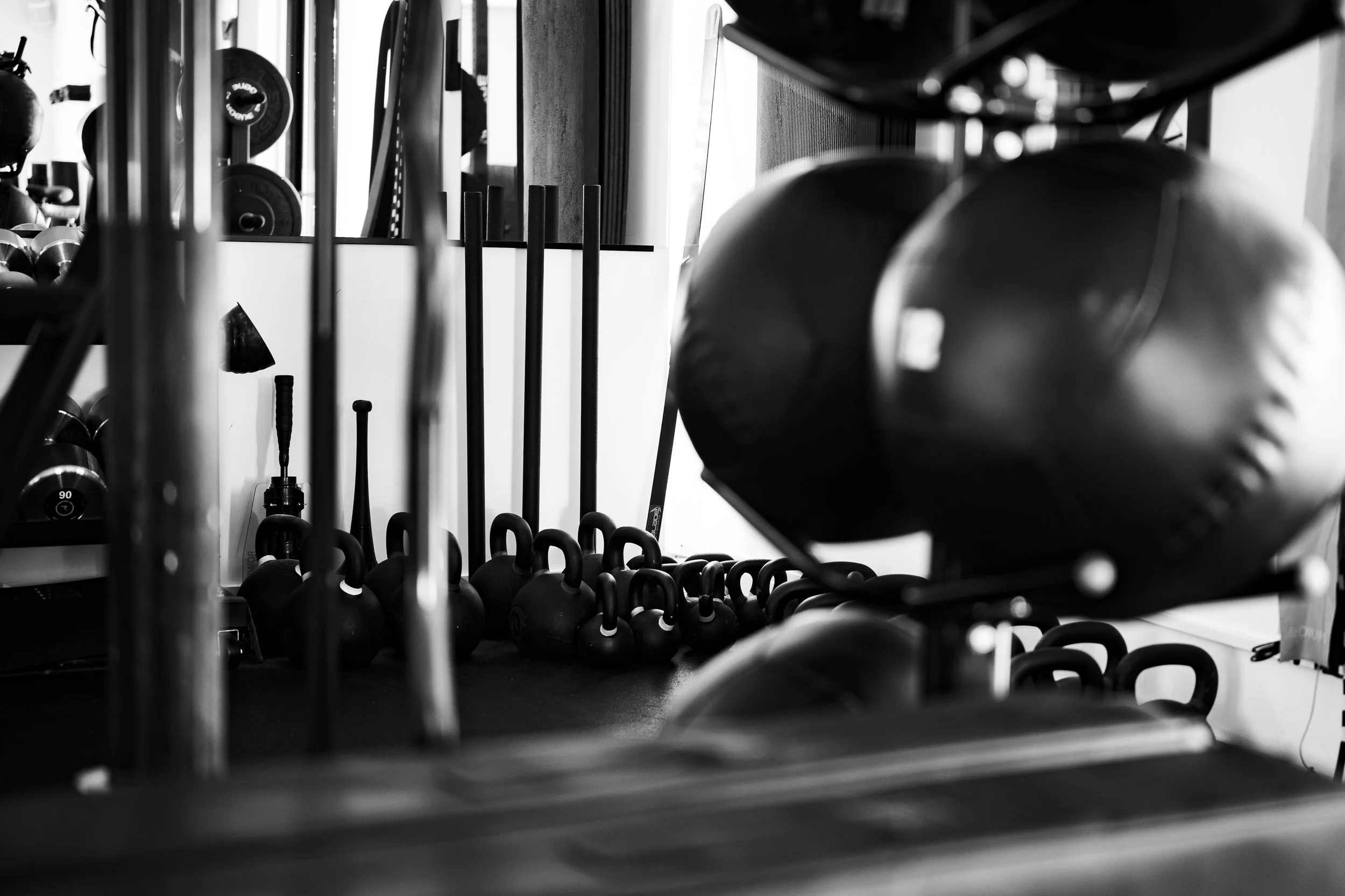 Black and white photo of various fitness equipment including kettlebells, medicine balls, and barbells, arranged on shelves and racks in a gym or workout space.