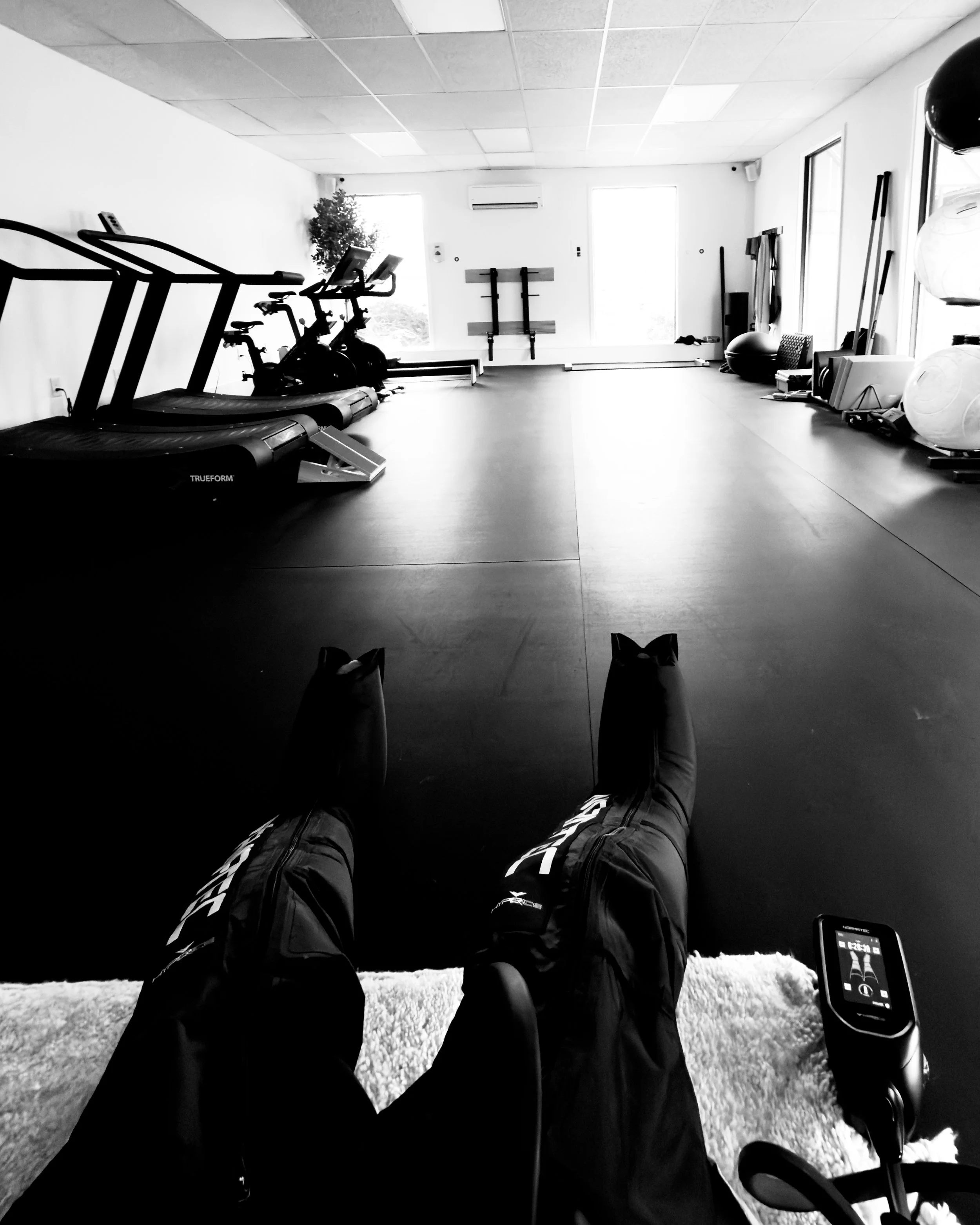 Person lying on the floor in a home gym, with their legs extended in the foreground, wearing sports shoes and workout pants. Fitness equipment such as a treadmill, exercise bikes, stability balls, and other workout gear are visible, with windows and 